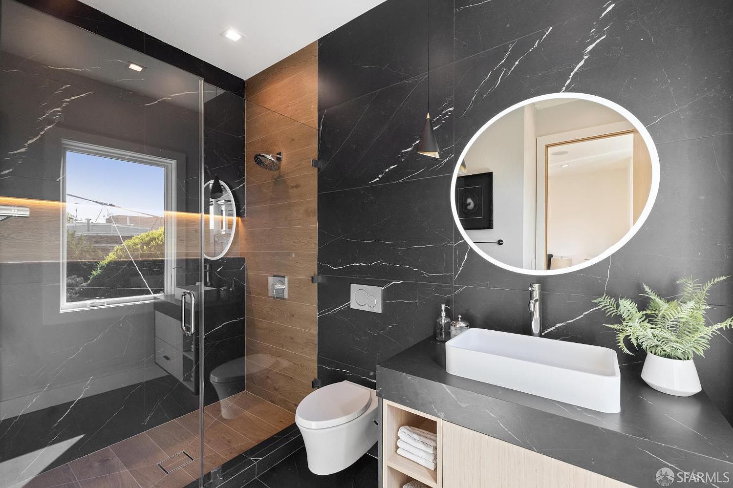 This is a modern primary bathroom featuring black marble walls with white veining, complemented by wood-paneled accents. The bathroom includes a glass-enclosed shower, a wall-mounted toilet, and a sleek white rectangular sink atop a dark countertop with light wood cabinetry. A round, illuminated mirror hangs above the sink, and a potted plant adds a touch of greenery.