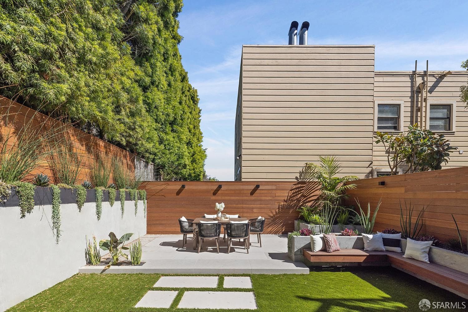 This image showcases a beautifully landscaped backyard, featuring a dining area with a table and chairs set on a concrete patio. A built-in bench with pillows provides additional seating, surrounded by lush greenery and a well-maintained lawn. The wooden fence and modern architectural details create a private and stylish outdoor space.