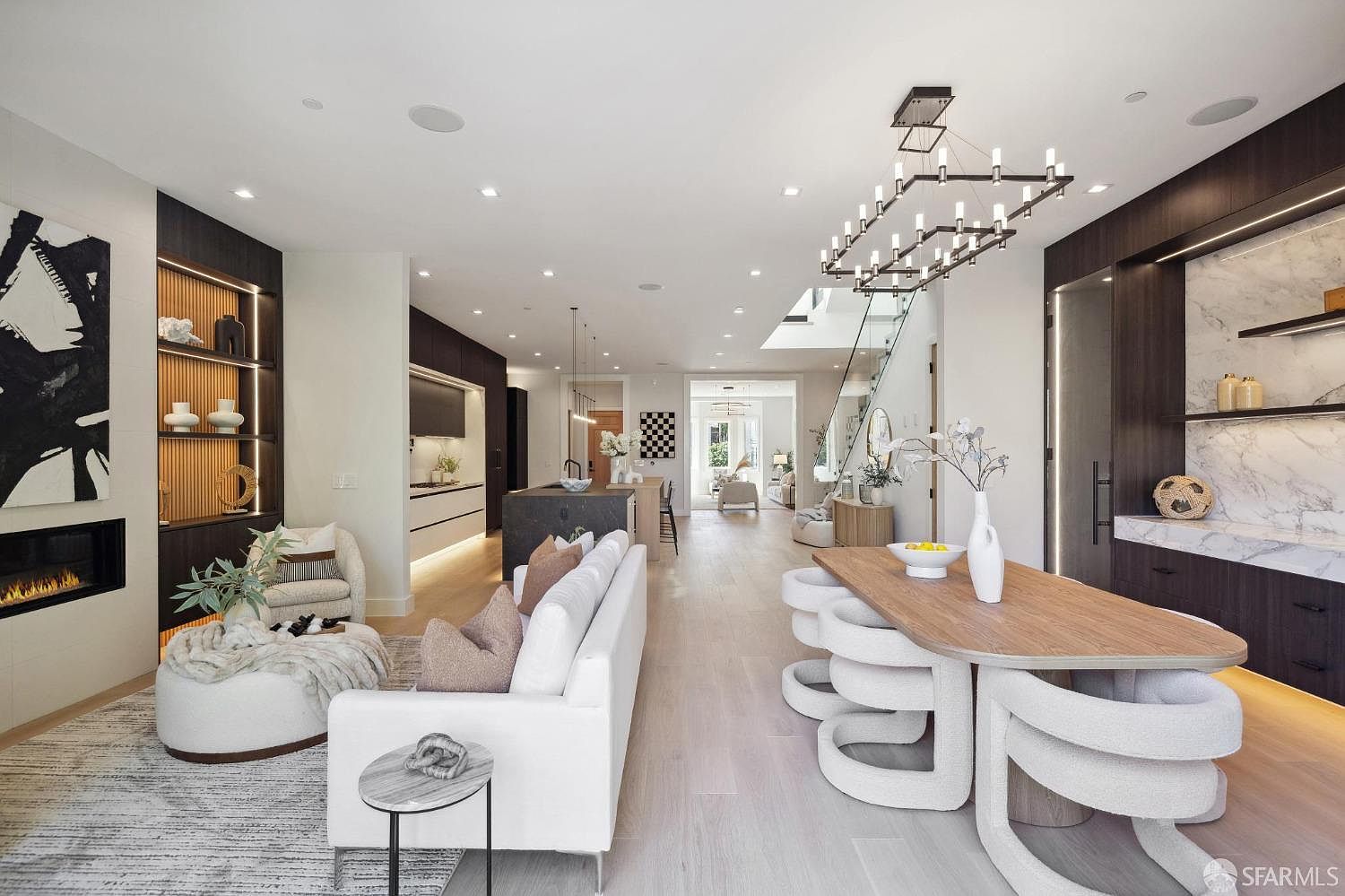 This is an interior shot of a modern living room, dining area, and kitchen. The living room features a fireplace, a white sofa, and a decorative rug. The dining area has a unique table with sculptural chairs, and the kitchen is equipped with sleek cabinetry and a central island. The open-concept design creates a spacious and inviting atmosphere, enhanced by natural light and contemporary lighting fixtures.