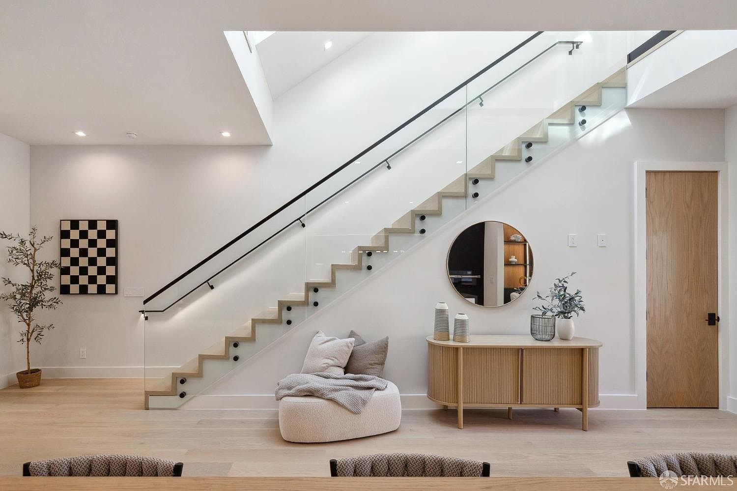 This interior shot showcases a modern hallway with a striking staircase featuring glass railings and wooden steps. A round mirror and a stylish console table add to the contemporary aesthetic. Natural light floods the space through a skylight, enhancing the bright and airy feel.