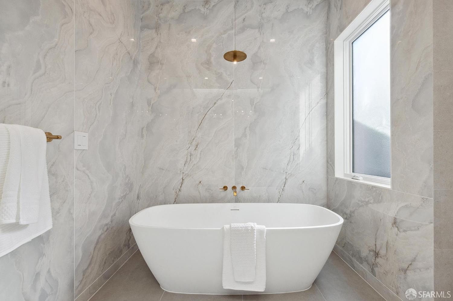 This is a luxurious primary bathroom featuring a freestanding white bathtub as the focal point. The walls are covered in elegant marble-like tiles, complemented by gold fixtures including a rainfall showerhead and faucet. A large window provides natural light, enhancing the spa-like atmosphere.