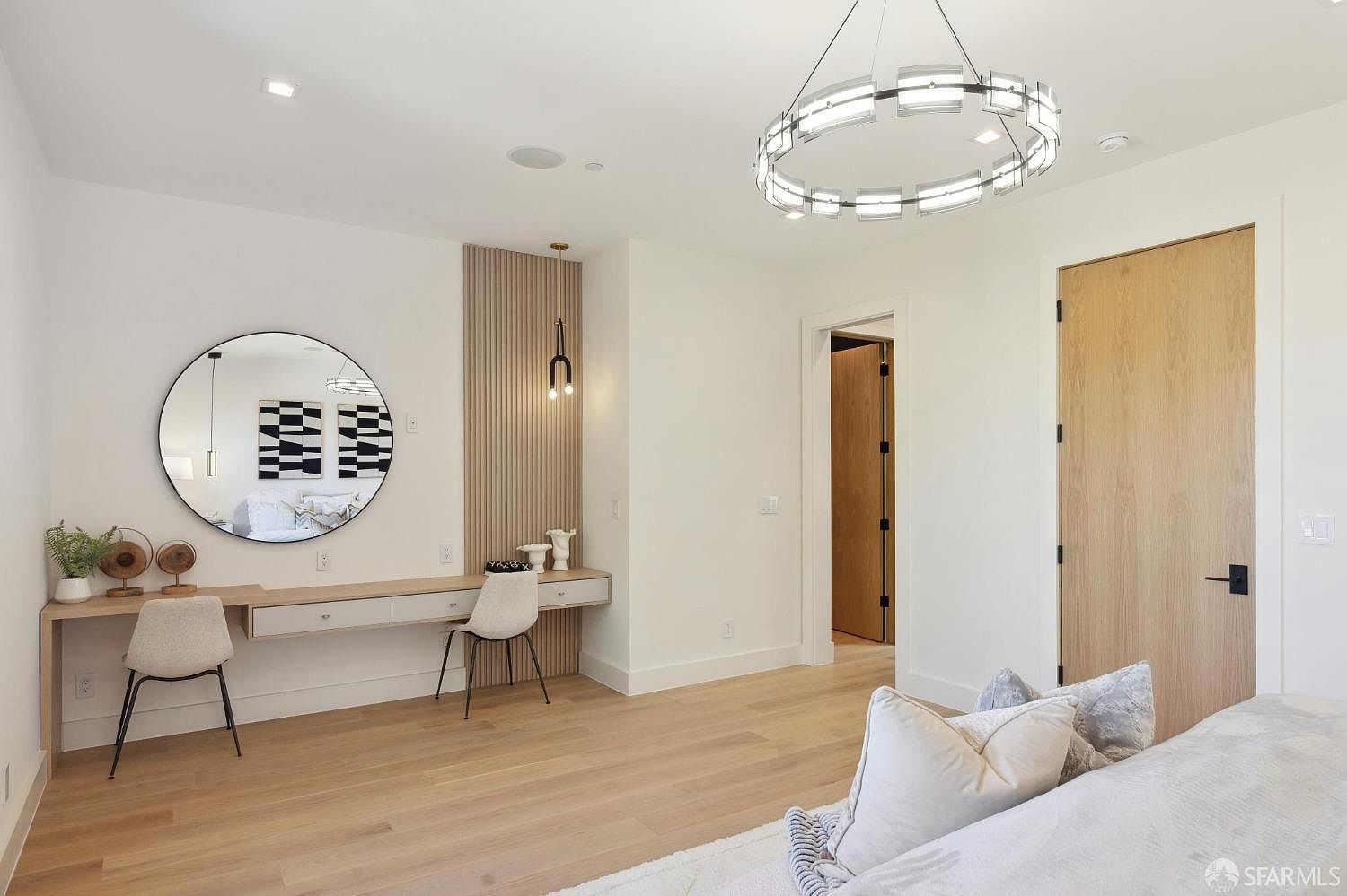 This is a bright and airy primary bedroom featuring a modern design with light wood flooring and white walls. A unique circular chandelier hangs from the ceiling, and a built-in vanity area with a round mirror and stylish chairs adds a touch of elegance. The room exudes a sense of calm and sophistication.