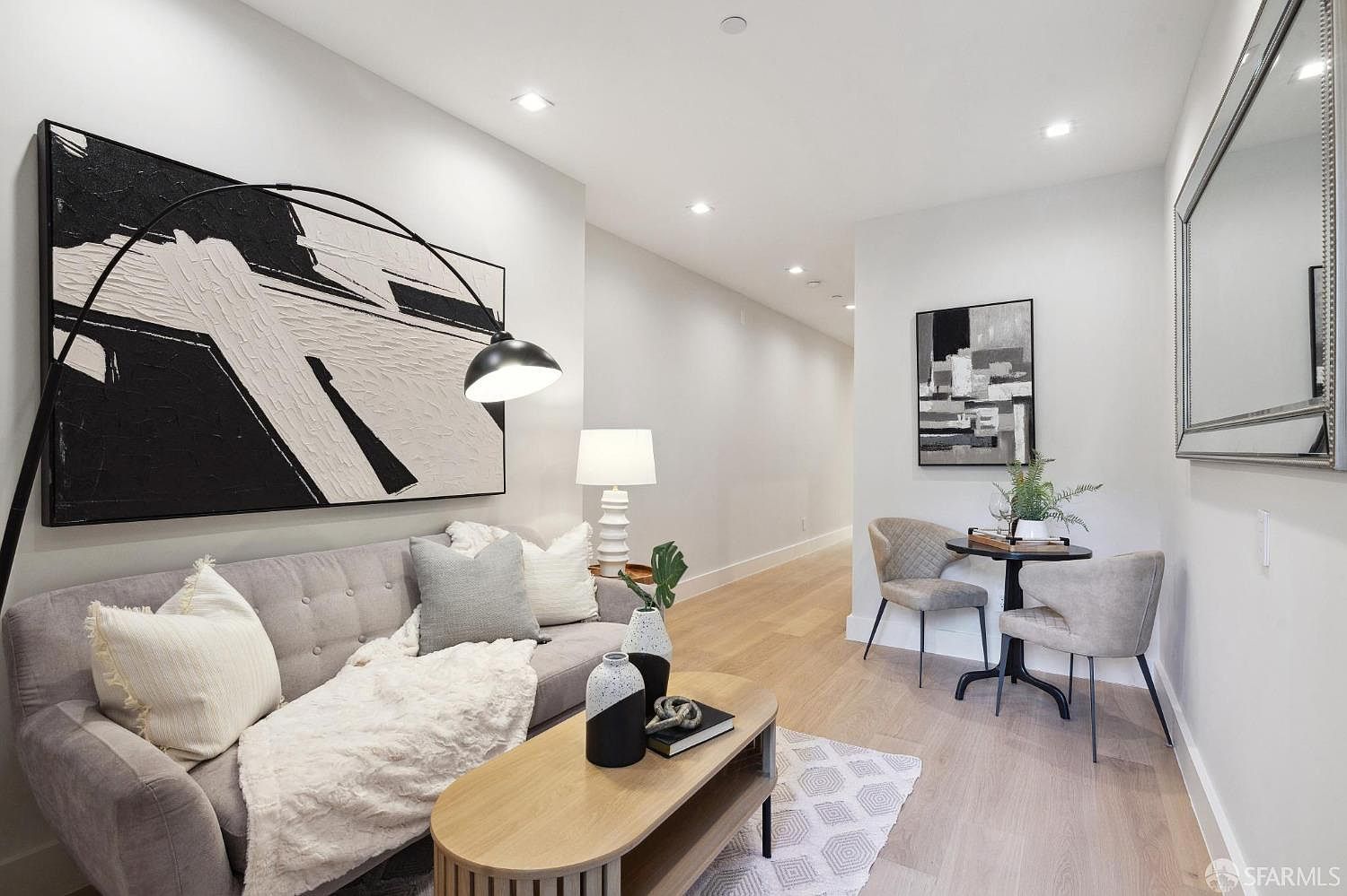 This is an interior shot of a living room featuring a gray sofa with decorative pillows and a throw blanket, a modern coffee table with vases and a book, and a large black and white abstract painting above the sofa. The room has light hardwood floors and neutral-colored walls, creating a bright and airy atmosphere. A small dining table with two chairs is visible in the background, adding to the functionality of the space.