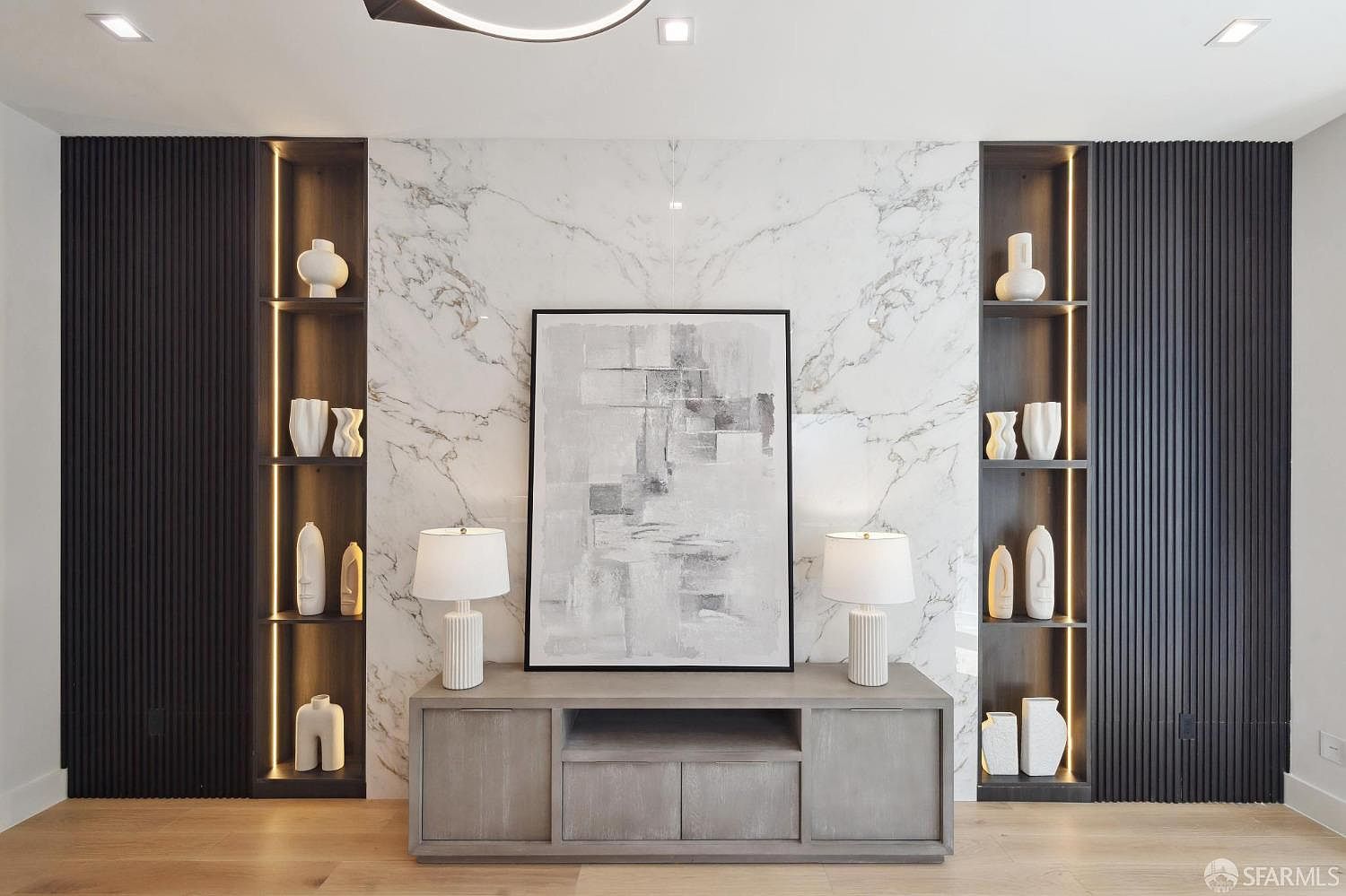 This interior shot showcases a modern living room with a focus on a stylish entertainment center. The center features dark wood paneling, marble accents, and illuminated shelving displaying decorative vases. A gray console table supports two lamps and a framed abstract painting, creating a sophisticated and inviting atmosphere.