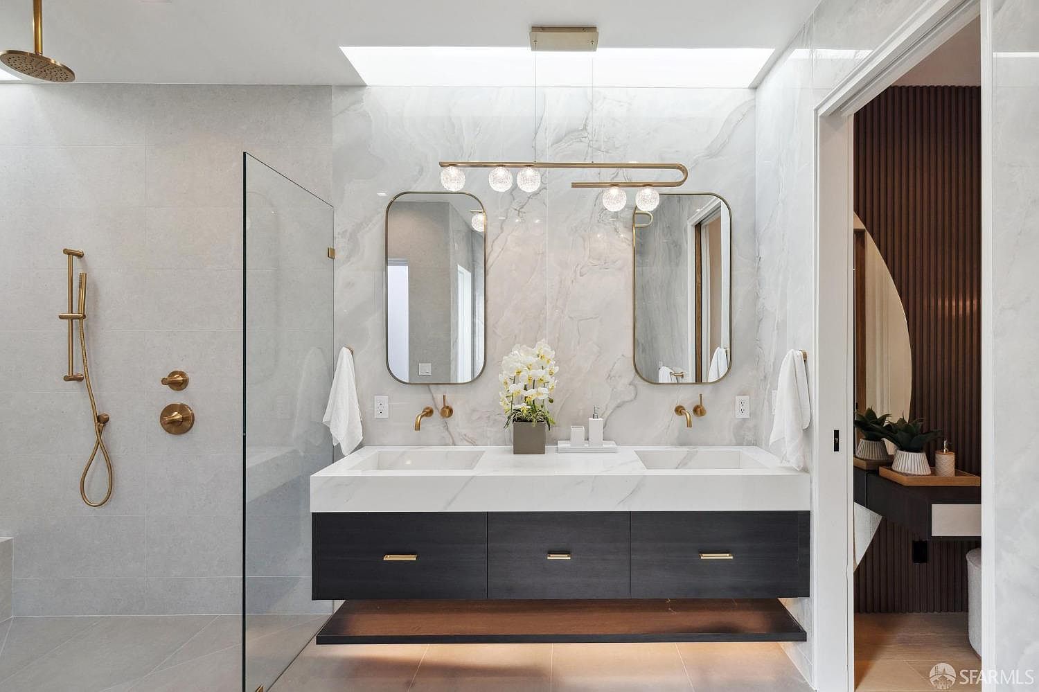 This is a luxurious primary bathroom featuring a double vanity with a white marble countertop and dark wood cabinetry. The walls are covered in marble tiles, and the space is illuminated by a modern light fixture and natural light from a skylight. Gold fixtures add a touch of elegance, and a glass-enclosed shower is visible to the left.