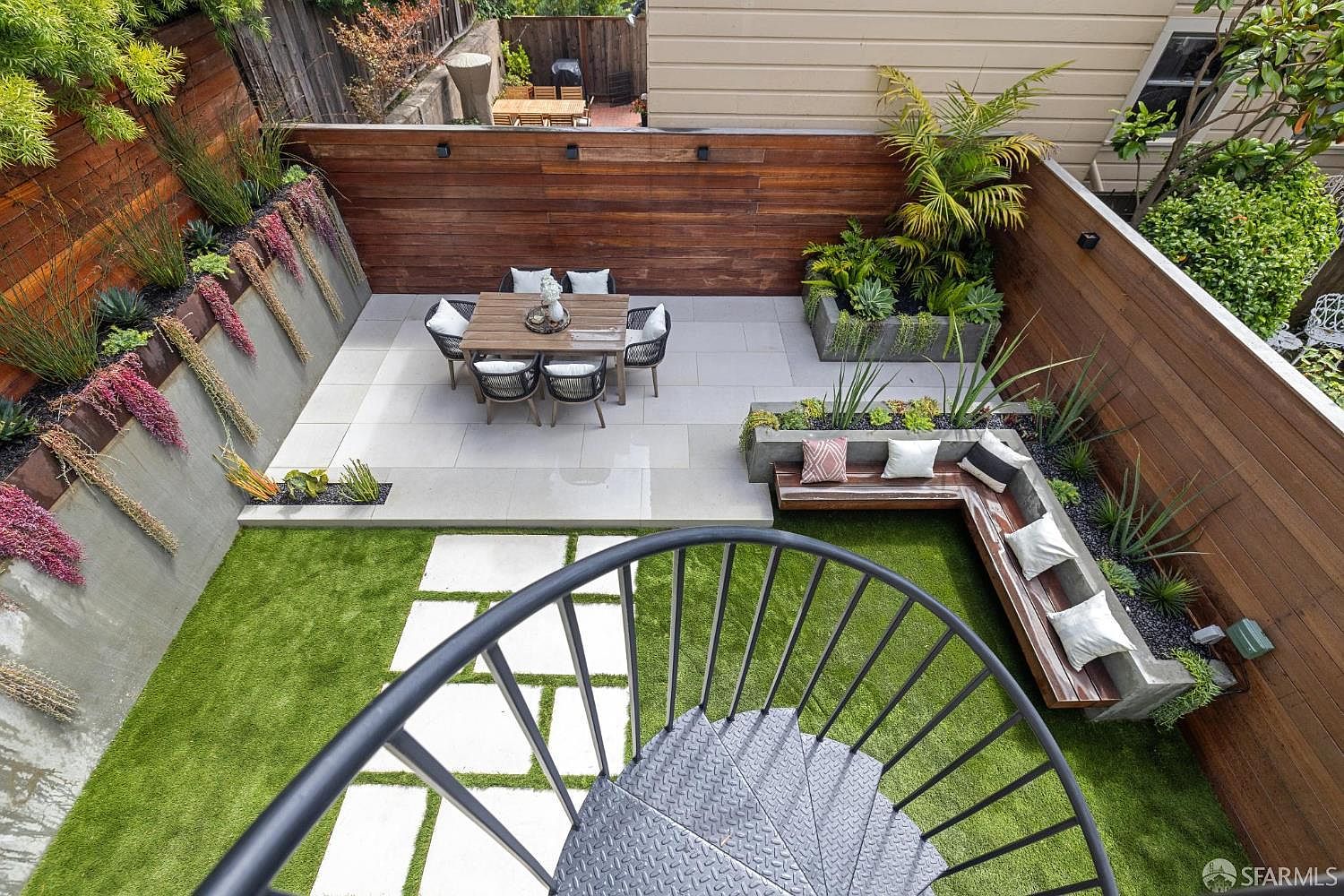 This image showcases a beautifully designed backyard garden. It features a dining area with a wooden table and chairs, a built-in bench with pillows, and lush greenery including plants and artificial turf. A spiral staircase leads down into the garden, adding a unique architectural element.
