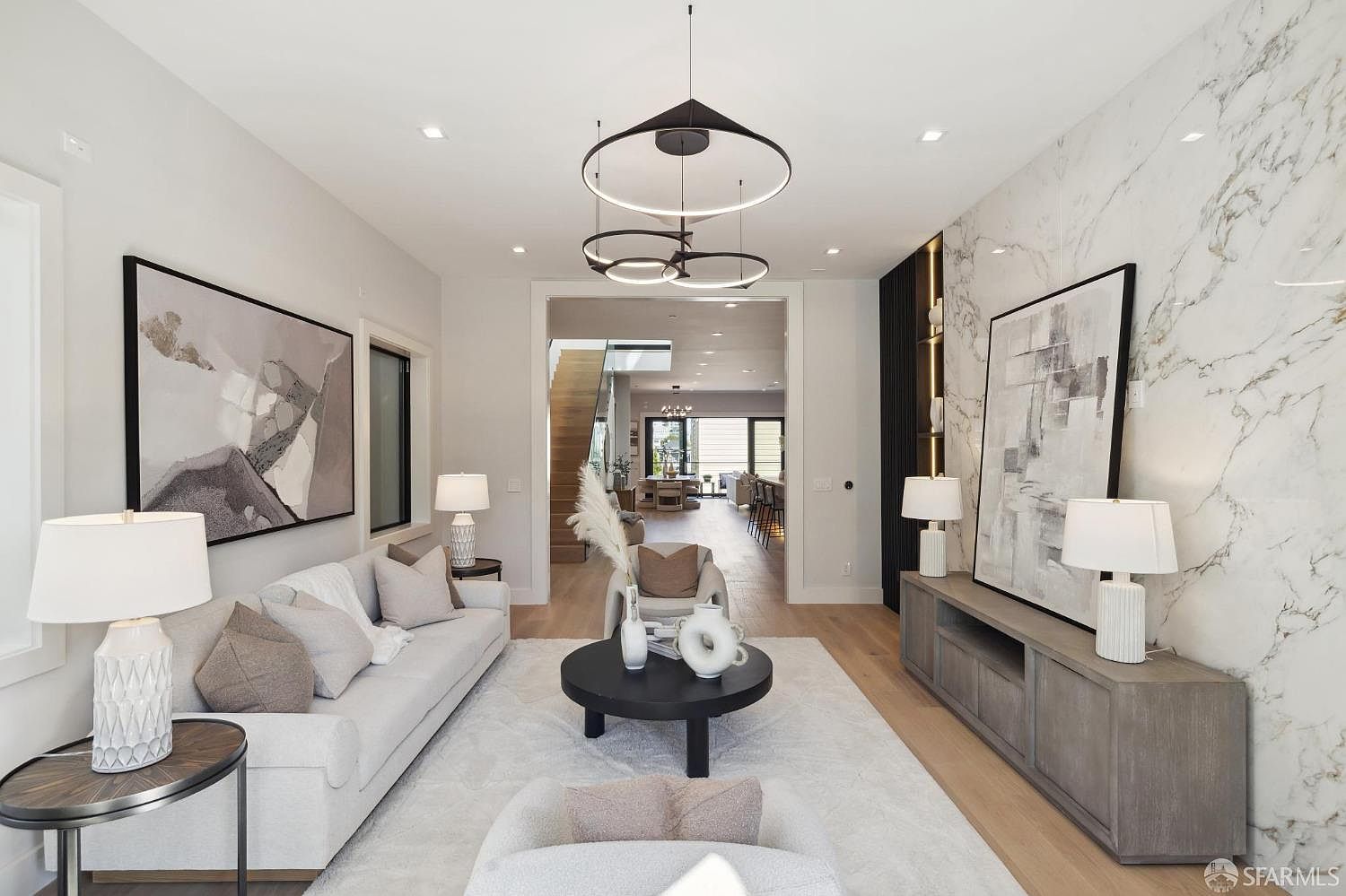This is a bright and airy living room featuring a neutral color palette with hardwood floors and a large area rug. The room is furnished with a light-colored sofa, armchairs, and a round coffee table. A striking marble accent wall and modern lighting fixtures add a touch of elegance, while artwork enhances the sophisticated ambiance.