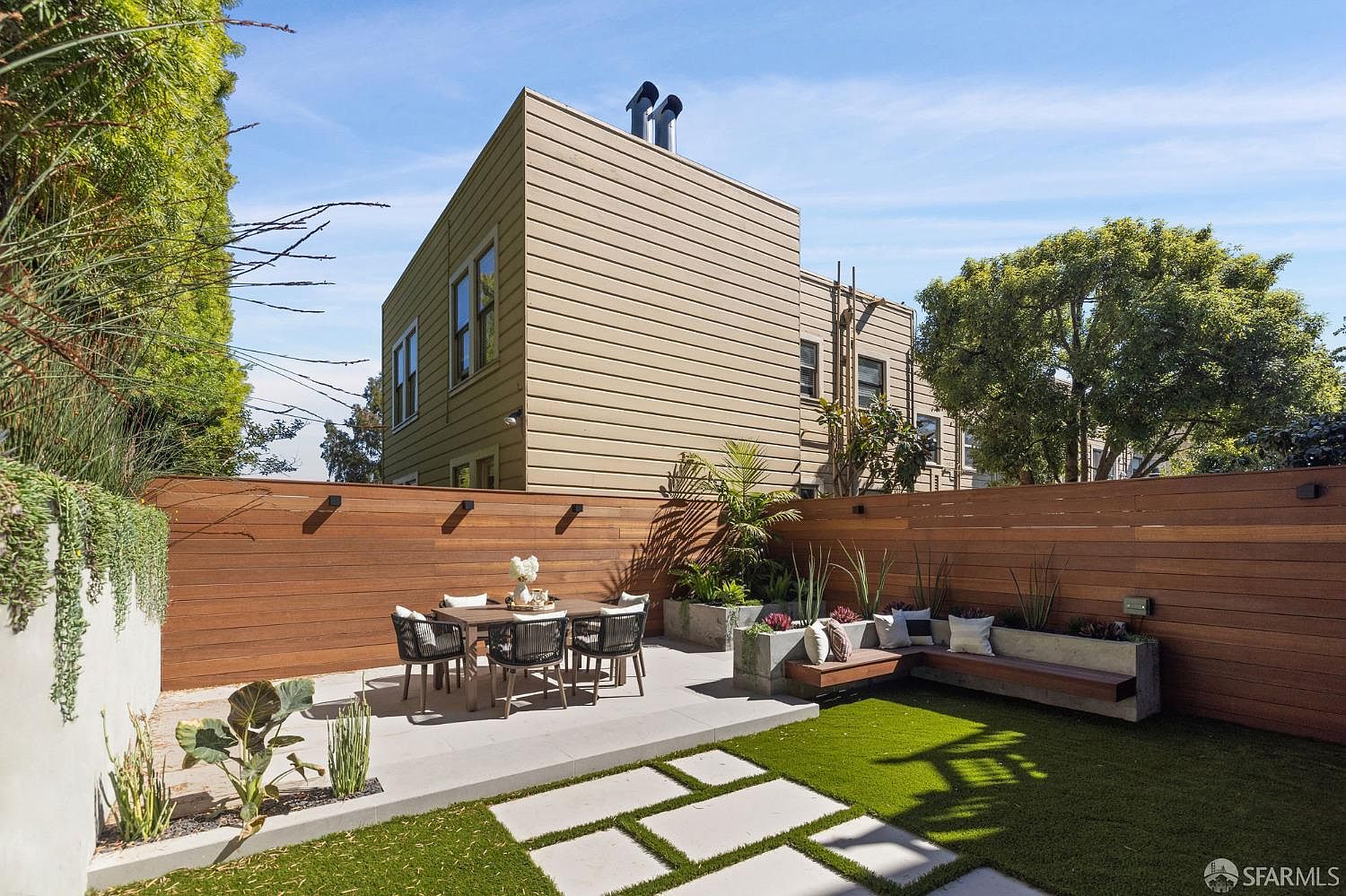 This is a well-designed backyard featuring a modern aesthetic. The space includes a dining area with a table and chairs, a built-in bench with cushions, and concrete planters with various plants. The yard is covered with artificial turf and features concrete pavers, all enclosed by a wooden fence, creating a private and stylish outdoor living area.