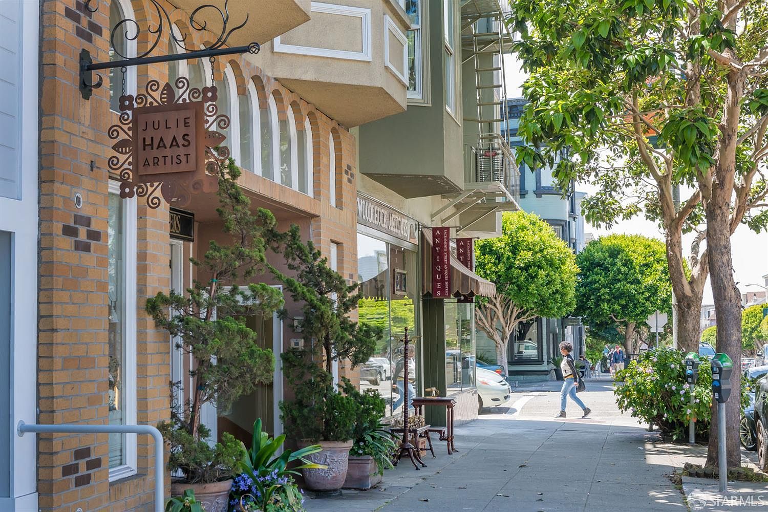 This is a street-level view of a commercial building featuring a brick facade and arched windows. Signage indicates the presence of an artist's studio and an antiques store. The sidewalk is lined with potted plants and trees, creating a pleasant urban atmosphere.