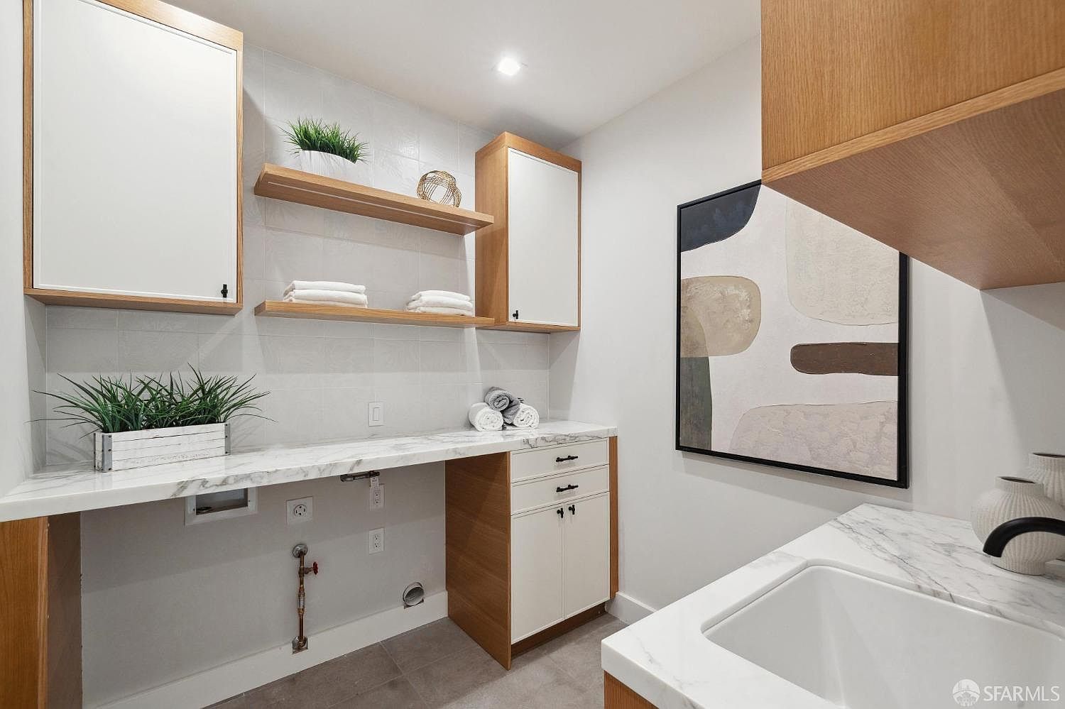 This is a well-organized laundry room featuring a marble countertop, white cabinets with wood trim, and open shelving. The room is brightly lit and includes decorative elements like plants and abstract art, creating a clean and modern aesthetic. The perspective is from a medium angle, showcasing the functional layout and design details.