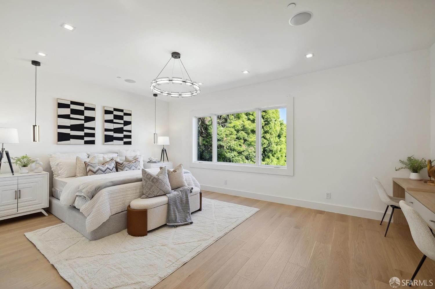 This is a bright and airy primary bedroom featuring a large window with a view of lush greenery. The room is decorated in a modern style with white walls, light wood floors, and contemporary lighting fixtures. The bed is neatly made with neutral-toned linens and decorative pillows, complemented by a stylish rug and a small seating area.