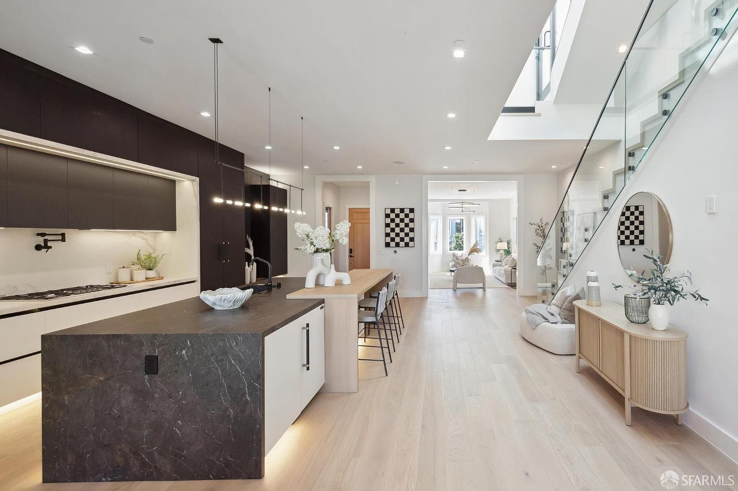 This is a bright and modern kitchen with a large island featuring a dark countertop and white cabinetry. The kitchen flows seamlessly into the living area, showcasing an open-concept design. A staircase with glass railings is visible, adding to the contemporary aesthetic of the home.