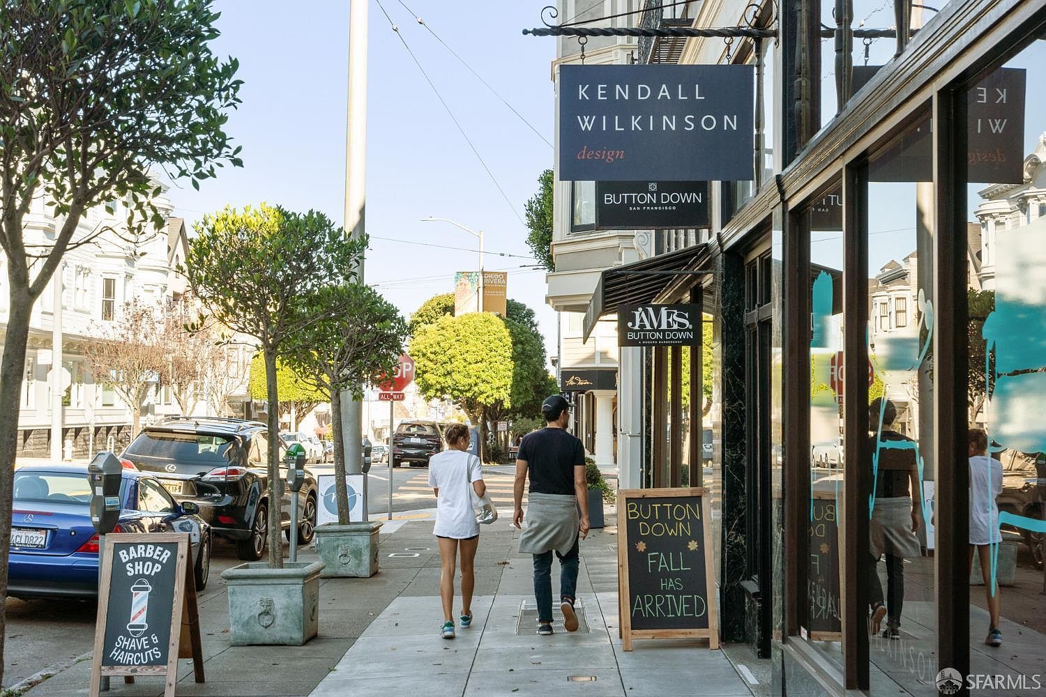 This image showcases a charming street view with a couple walking along the sidewalk, lined with trees and well-maintained planters. Several businesses are visible, including a barber shop and a design studio, indicated by prominent signage. The overall impression is a vibrant and walkable neighborhood with a mix of commercial and residential elements.