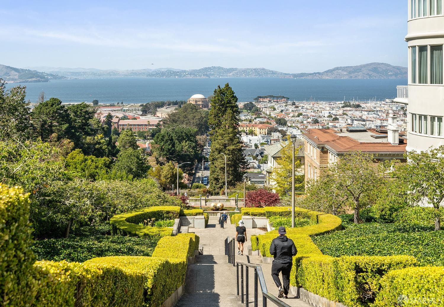 This image showcases a beautifully landscaped garden with meticulously trimmed hedges lining a descending staircase. In the background, a panoramic view of the city and bay adds significant value, highlighting the property's prime location. The presence of people walking down the stairs suggests accessibility and usability of the outdoor space.