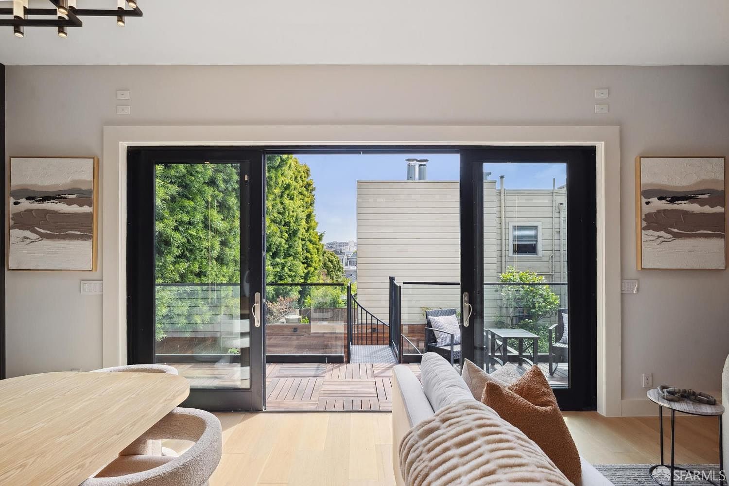 This interior shot showcases a bright living room with a seamless transition to an outdoor deck through large sliding glass doors. The room features modern artwork, light wood flooring, and a comfortable-looking sofa, creating an inviting and airy atmosphere. The outdoor deck offers additional living space and a view of the surrounding neighborhood.