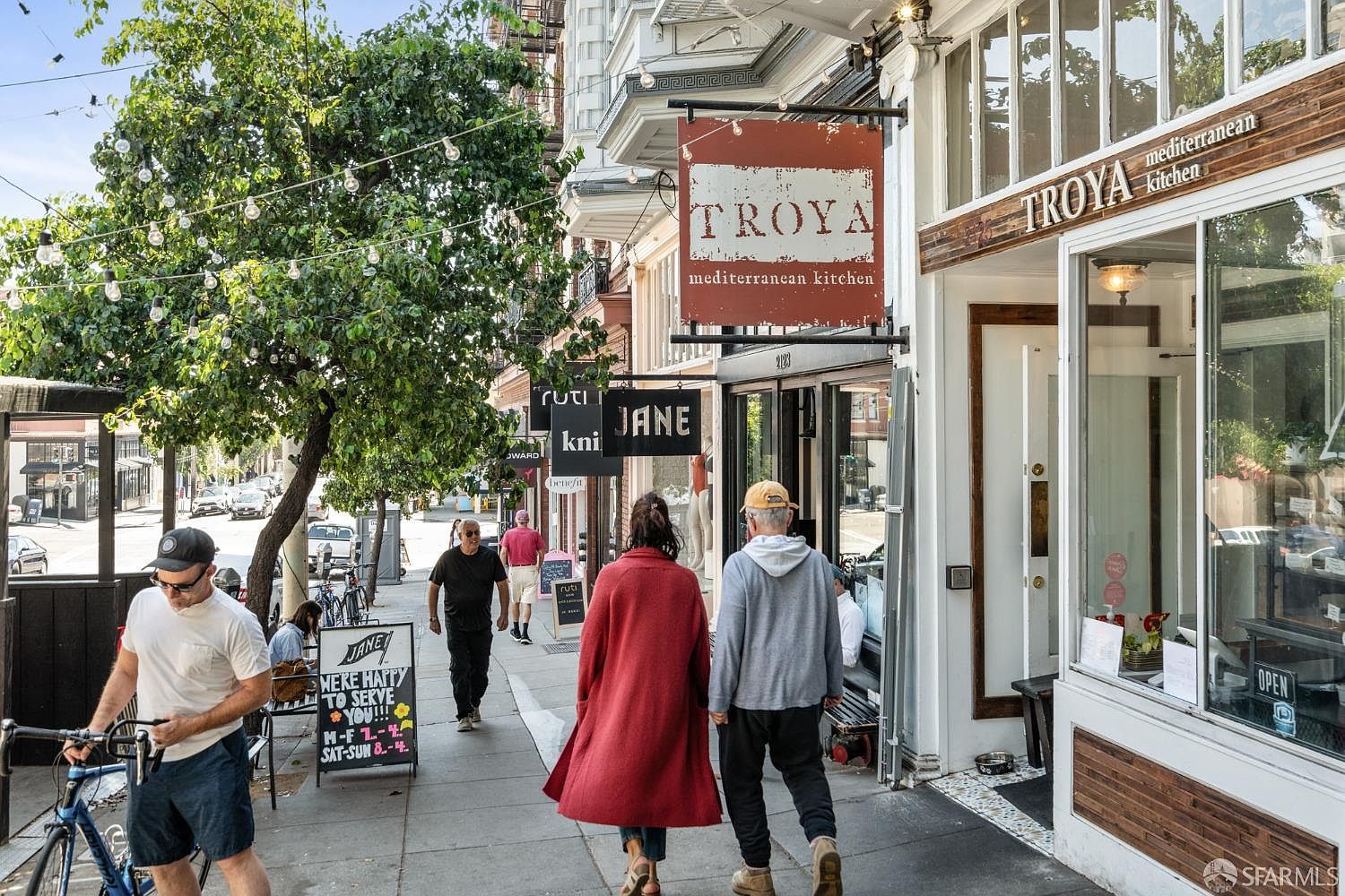 This image showcases the front exterior of a building with commercial spaces, including the "Troya Mediterranean Kitchen." Pedestrians stroll along the sidewalk, adding a sense of urban activity. The architectural style features classic building facades with detailed trim and large windows, creating an inviting street presence.
