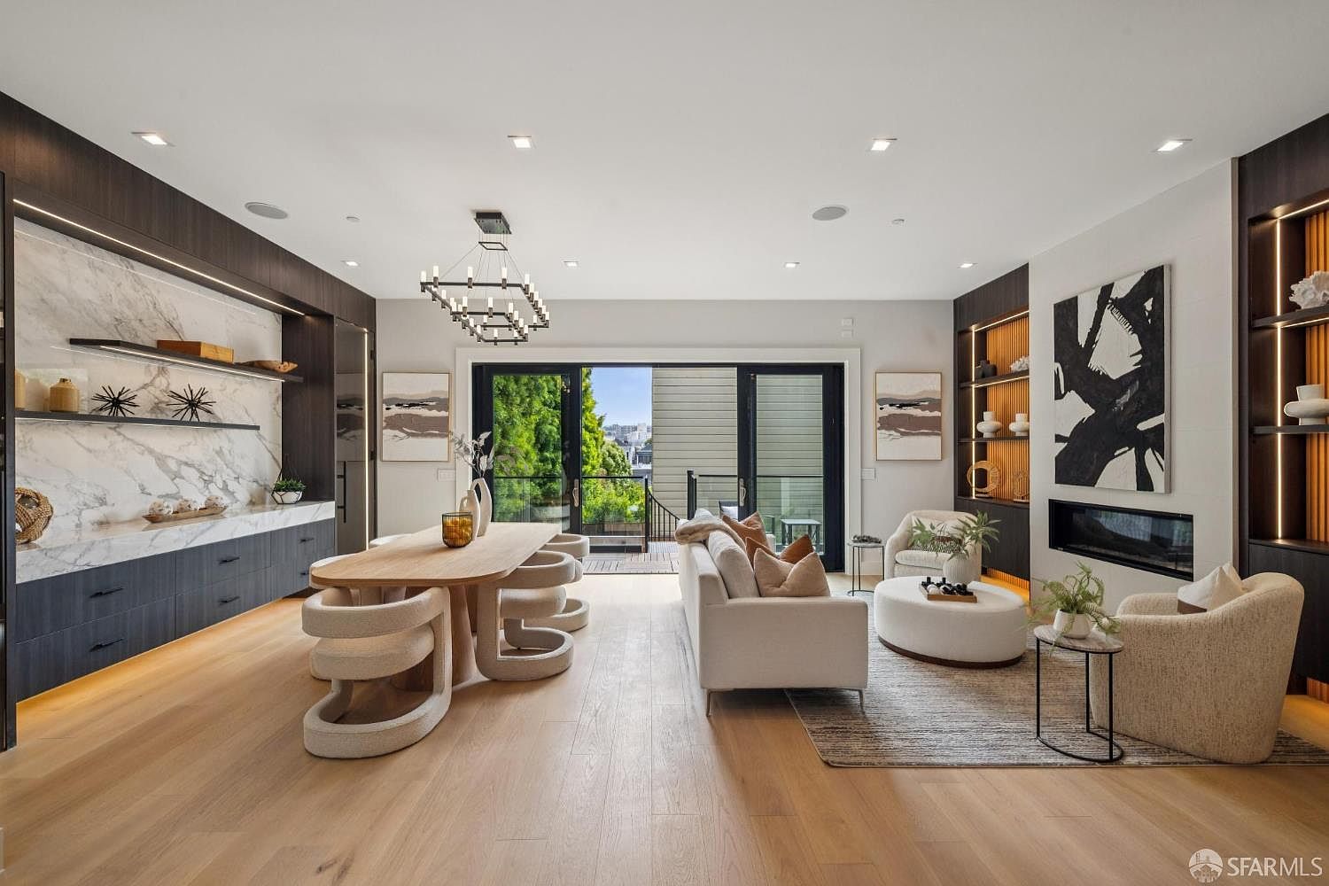 This is a bright and modern living and dining area featuring hardwood floors, a contemporary dining table with unique chairs, and a comfortable living space with a white sofa and ottoman. The room is well-lit with natural light from the sliding glass doors leading to a balcony, and it includes stylish built-in shelving and a fireplace, creating a sophisticated and inviting atmosphere.
