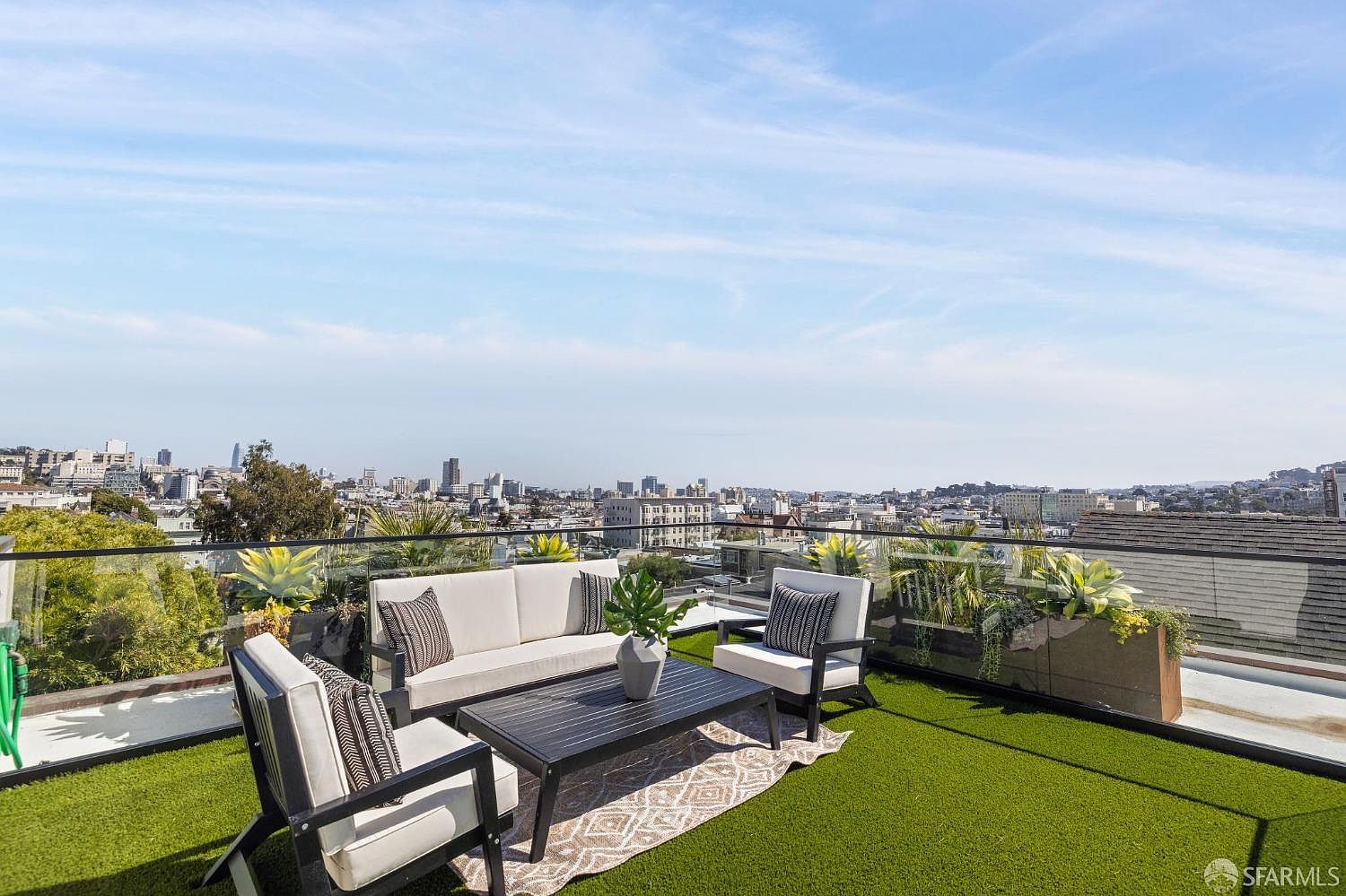 This image showcases a stylish rooftop patio with a stunning city view. The patio features modern outdoor furniture, including a sofa, chairs, and a coffee table, arranged on artificial turf. A glass railing provides an unobstructed view of the cityscape, enhancing the property's appeal for outdoor living and entertaining.