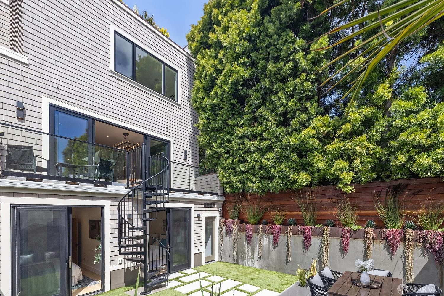 This is a rear view of a multi-story house with gray wood siding. A black spiral staircase connects the upper balcony to the ground level patio. The patio features a small lawn area with stone pavers, a wooden fence with planter boxes, and an outdoor dining set, creating a cozy and private backyard space.