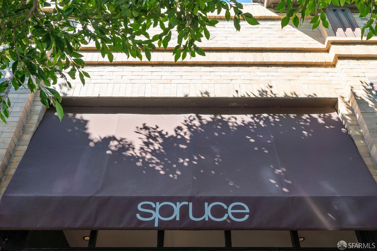 The image showcases the exterior entryway of a building, featuring a brown awning with the word "spruce" printed on it. The building's facade is constructed of light-colored bricks, and the scene is partially shaded by tree branches with green leaves, creating dappled shadows on the awning. The perspective is a medium shot, focusing on the entrance and highlighting the building's architectural details.