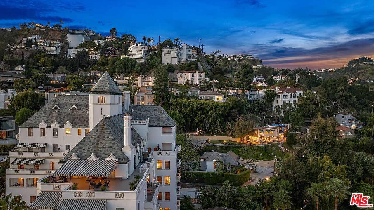 This aerial view showcases a grand, multi-story building with a distinctive turret and gray-tiled roof, nestled among lush greenery and other hillside homes. The building features multiple balconies and terraces, suggesting outdoor living spaces, while the surrounding landscape adds to the property's privacy and appeal. The image captures a serene, upscale residential setting at dusk.