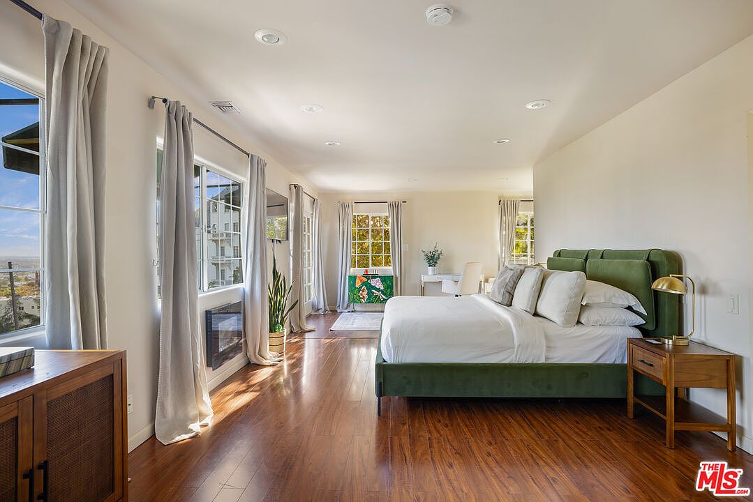This is a bright and spacious primary bedroom featuring hardwood floors, neutral-toned walls, and large windows with light gray curtains. The room includes a green upholstered bed with white linens, a wooden nightstand with a gold lamp, and a decorative fireplace. A desk area is visible in the background, creating a versatile and inviting space.