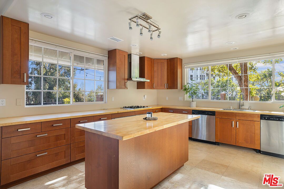 This is a well-lit kitchen featuring wooden cabinetry, stainless steel appliances, and a central island with a light-colored countertop. Large windows provide ample natural light and views of the outdoors. The kitchen has a warm and inviting feel, with a focus on functionality and natural light.