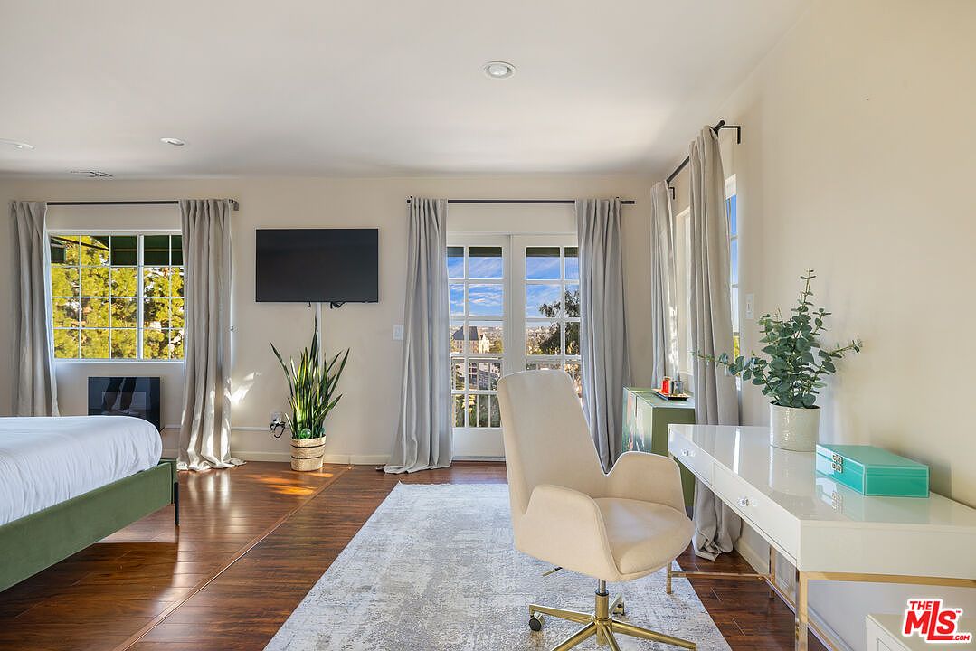 This primary bedroom features hardwood floors, a large window with natural light, and a flat-screen TV mounted on the wall. A desk with a modern chair creates a functional workspace, while a bed with a green frame adds a touch of color. The room is spacious and well-lit, offering a comfortable and stylish living space.