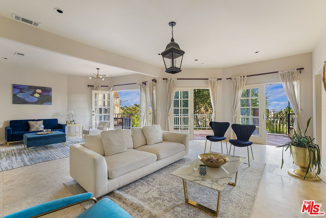 This is a bright and airy living room featuring a neutral color palette with pops of blue. The room is furnished with a large white sofa, a blue sofa, and a marble-topped coffee table. French doors lead to a balcony, providing natural light and views of the outdoors.