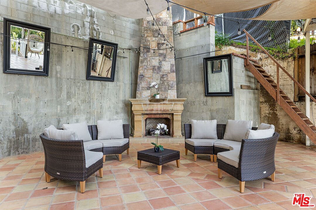 This image showcases an outdoor patio area designed for relaxation and entertainment. The space features comfortable wicker seating arranged around a central coffee table, creating an inviting conversation area. A stone fireplace adds a focal point, while string lights overhead provide ambient lighting, enhancing the cozy atmosphere.