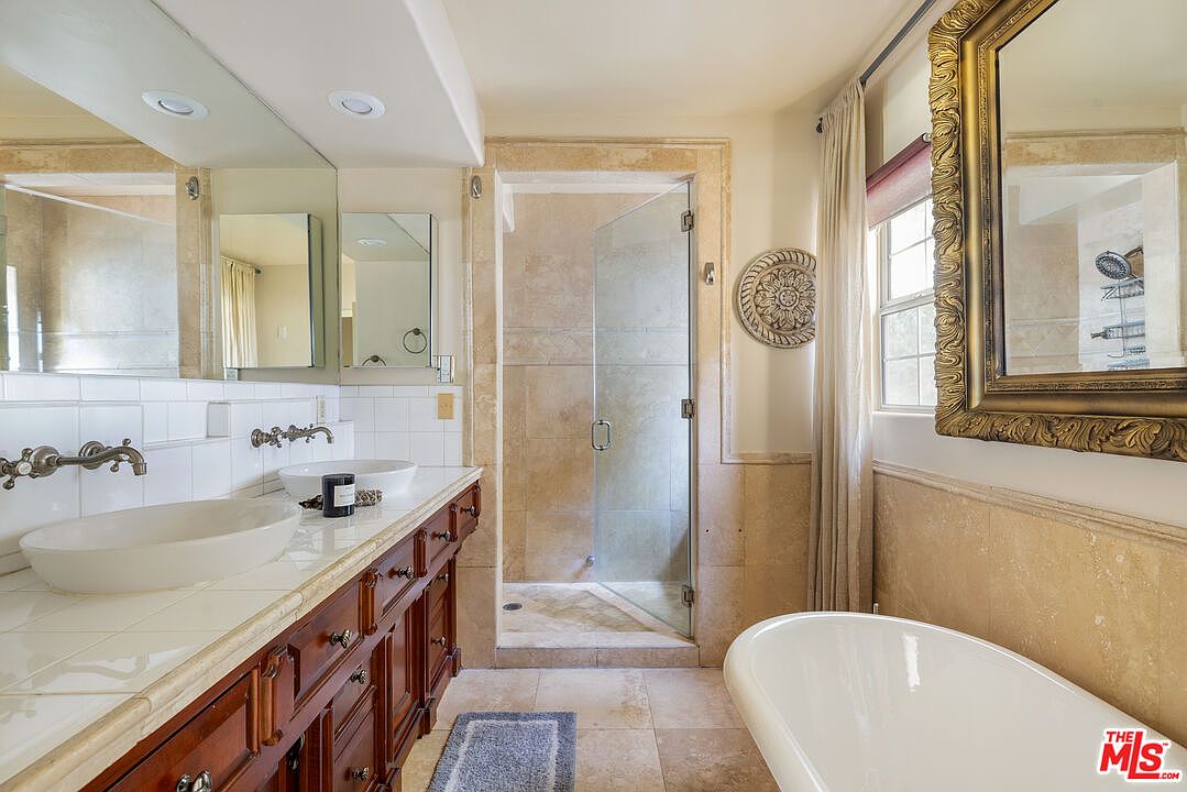 This primary bathroom features a double vanity with vessel sinks, a tiled countertop, and dark wood cabinetry. A glass-enclosed shower is visible in the background, and a freestanding bathtub is positioned near a window with a decorative mirror. The bathroom has a luxurious and classic design with neutral tones and tile flooring.