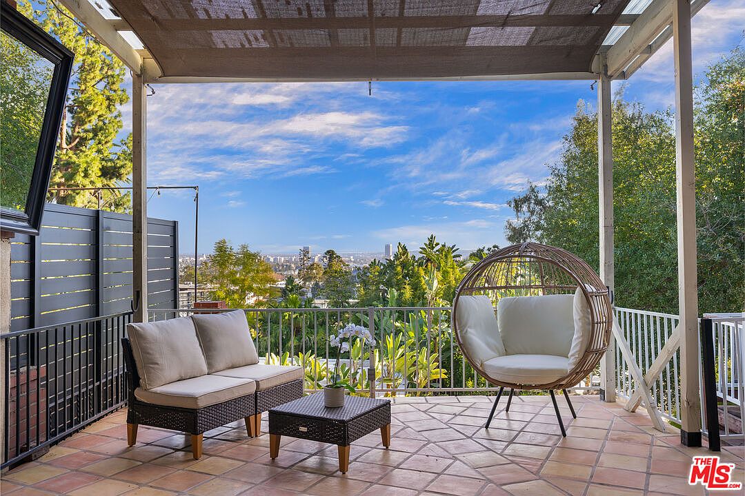This image showcases a charming patio or balcony area, furnished with a wicker loveseat, a unique round chair, and a small table, all arranged on a tiled floor. The space is partially covered by a pergola-like structure, offering shade, while the railing provides safety and a clear view of the cityscape and lush greenery in the background. The overall impression is one of relaxation and outdoor living.
