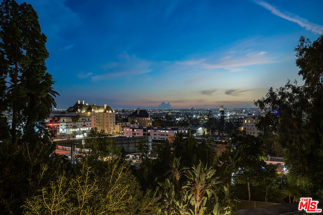 This aerial view showcases a cityscape at twilight, with illuminated buildings and a vibrant blue sky fading into the horizon. Lush trees frame the foreground, adding a touch of nature to the urban panorama. The image captures a dramatic and appealing perspective of the property's location and surrounding environment.