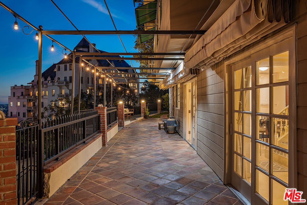 This image showcases a charming patio or balcony area, featuring a tiled floor and a metal pergola adorned with string lights, creating a warm and inviting ambiance. The space is bordered by a brick and iron railing, offering a view of a grand building in the background. The patio is adjacent to a building with a covered awning and glass doors, suggesting seamless indoor-outdoor living.
