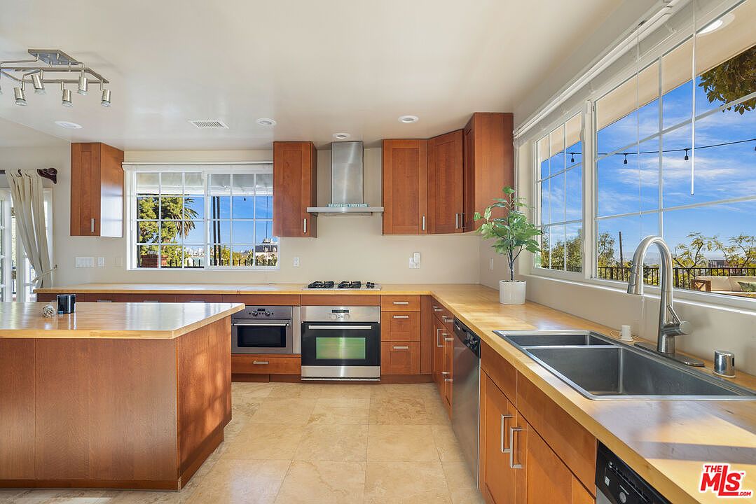 This is a well-lit kitchen featuring wooden cabinetry and countertops, stainless steel appliances, and a central island. Large windows provide natural light and views of the outdoors. The kitchen has a warm and inviting feel, perfect for cooking and entertaining.