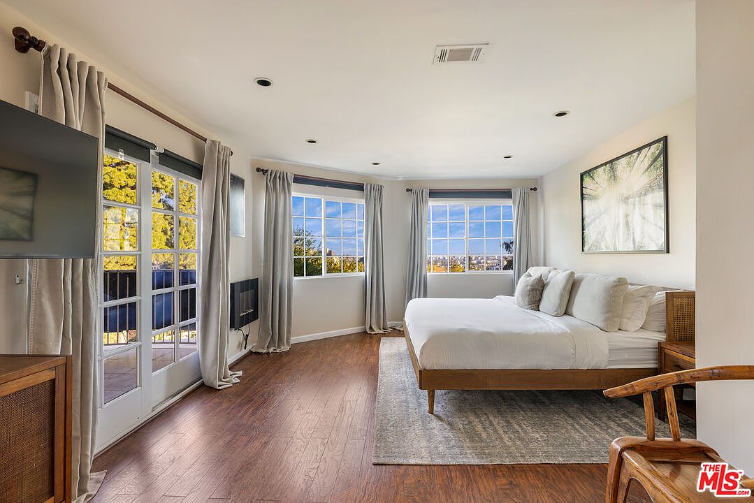 This is a bright and airy primary bedroom featuring hardwood floors, multiple windows with views, and a neutral color palette. The room is furnished with a bed dressed in white linens, a wooden side table, and a modern art piece on the wall. The overall impression is clean, comfortable, and inviting.