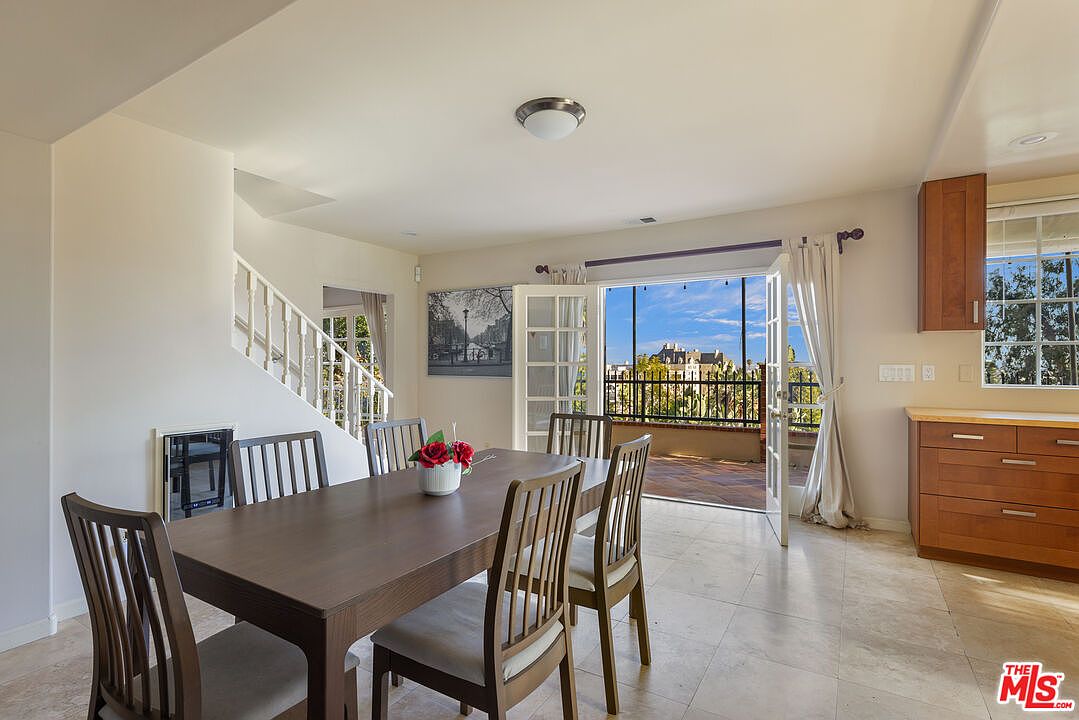 This dining room features a dark wood dining table with seating for six, set on a light tile floor. French doors open to a balcony, providing natural light and a view. A staircase is visible to the left, and built-in cabinetry is on the right, creating a functional and inviting space.