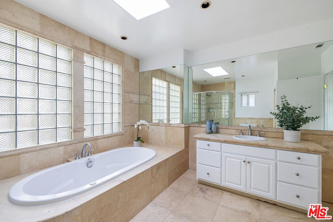 This primary bathroom features a large soaking tub surrounded by beige tile, complemented by two glass block windows for natural light. A vanity with white cabinets and a beige countertop sits adjacent to the tub, topped with a large mirror and a potted plant. The overall impression is a clean and luxurious space.