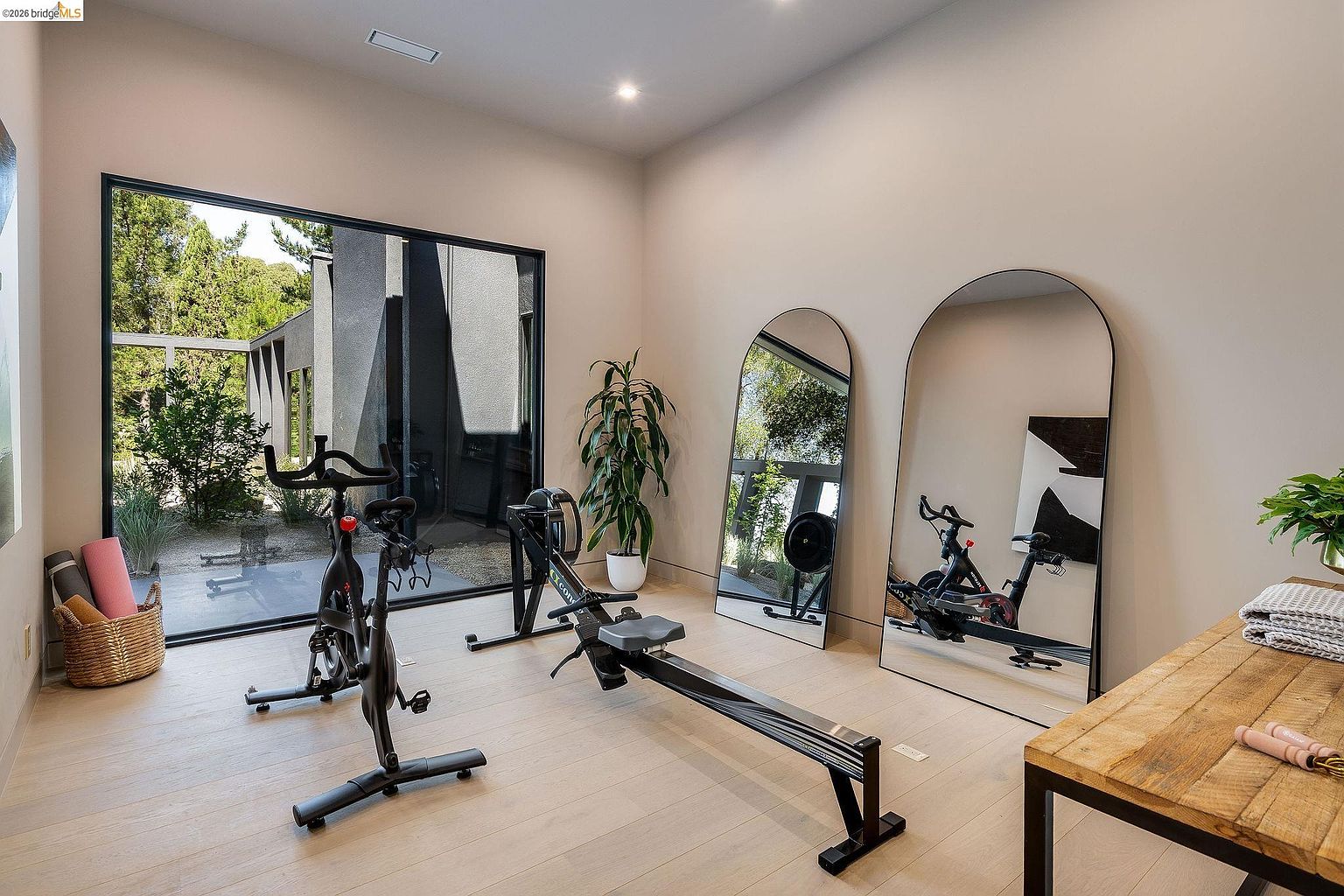 This modern home gym features light wood flooring, neutral walls, and a large floor-to-ceiling window that provides a bright, airy atmosphere with views of the outdoor landscape. The space is equipped with a stationary bike, a rowing machine, and two large arched mirrors that enhance the room's depth and natural light. A rustic wooden console table in the foreground adds a touch of warmth to the clean, minimalist aesthetic.