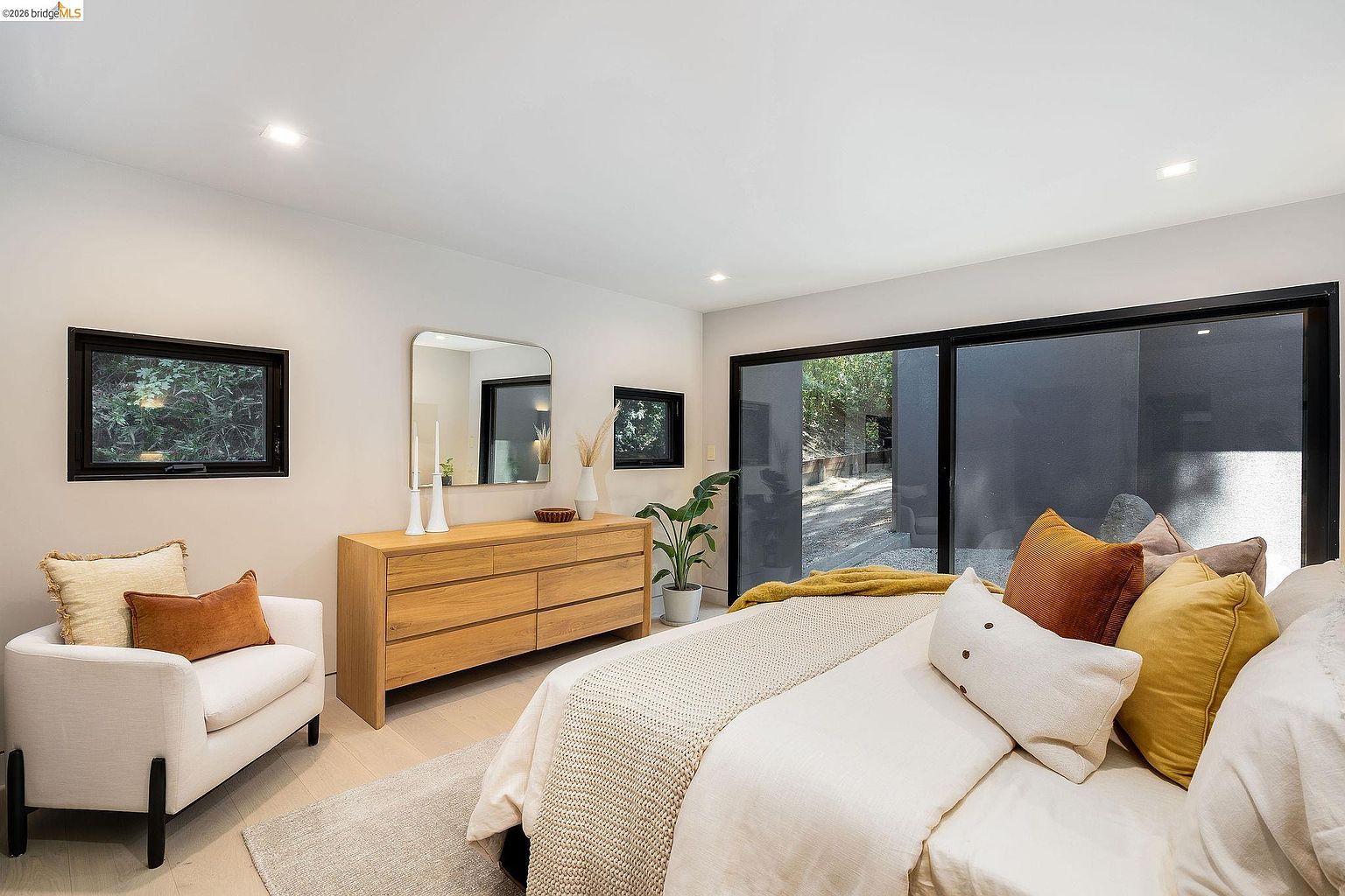 This serene primary bedroom features a contemporary design with a neutral color palette, accented by warm wood tones and rich textures. A large sliding glass door provides abundant natural light and a seamless connection to the outdoor space, while a stylish dresser and armchair complete the inviting, minimalist aesthetic. The room is thoughtfully staged to emphasize comfort and modern elegance.