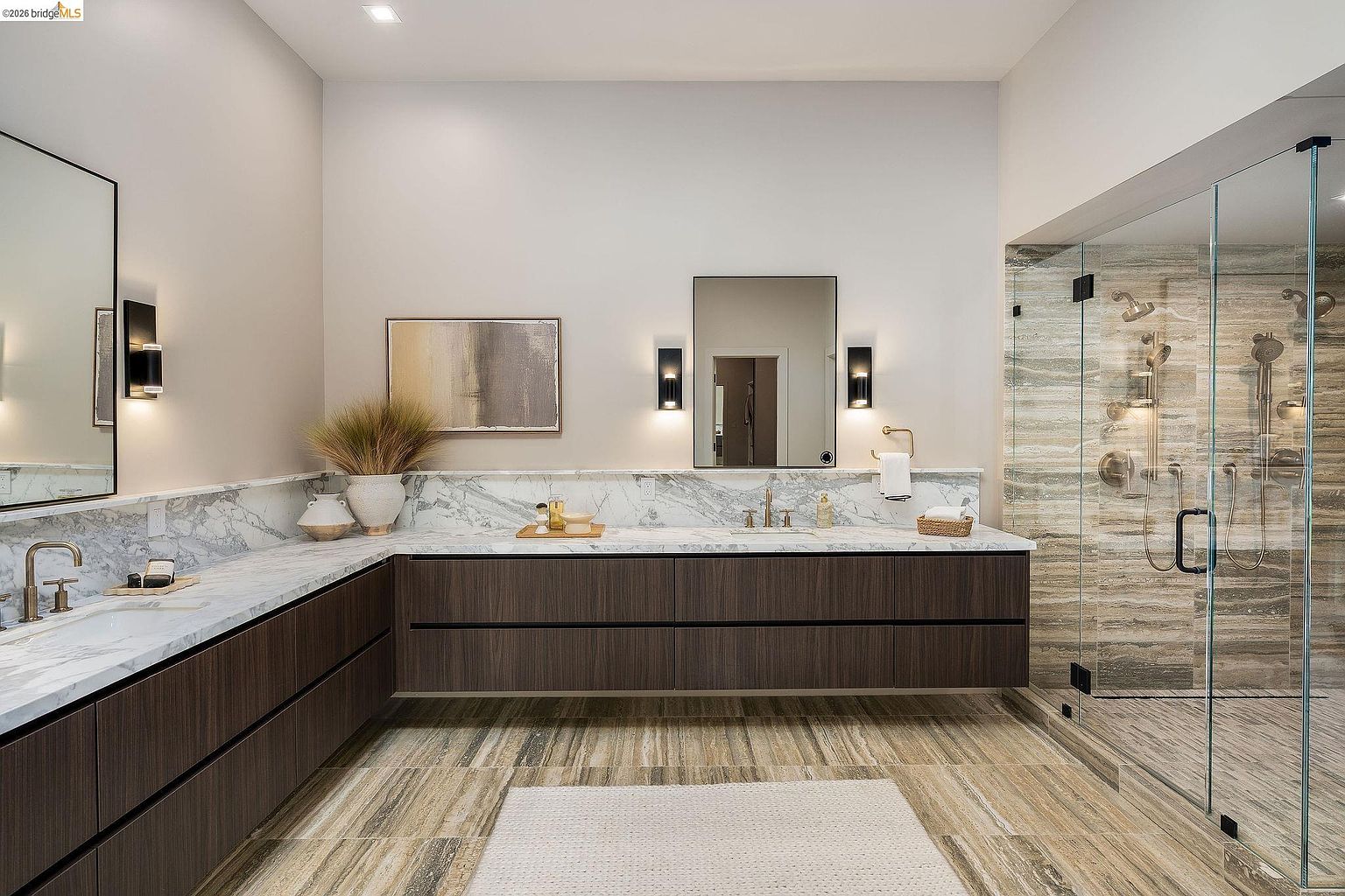 This luxurious primary bathroom features a sleek, L-shaped floating vanity with dark wood cabinetry and a striking white marble countertop. A spacious walk-in shower with textured stone tile walls and glass enclosures anchors the right side of the room, while elegant wall sconces and mirrors add a sophisticated touch. The overall aesthetic is modern and high-end, creating a serene, spa-like atmosphere.