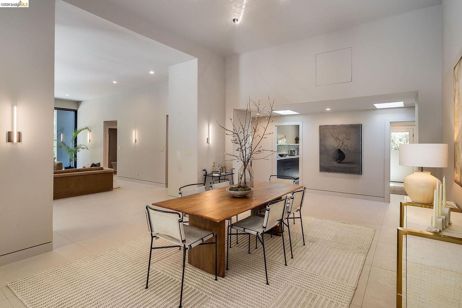 This elegant dining room features a warm wooden table surrounded by modern, minimalist chairs, set atop a textured area rug. The high ceilings and neutral color palette create an airy, sophisticated atmosphere, while a large piece of wall art and a decorative lamp add character to the space. The perspective captures the open-concept flow, looking past the dining area toward a bright living room and a glimpse of a kitchen in the background.