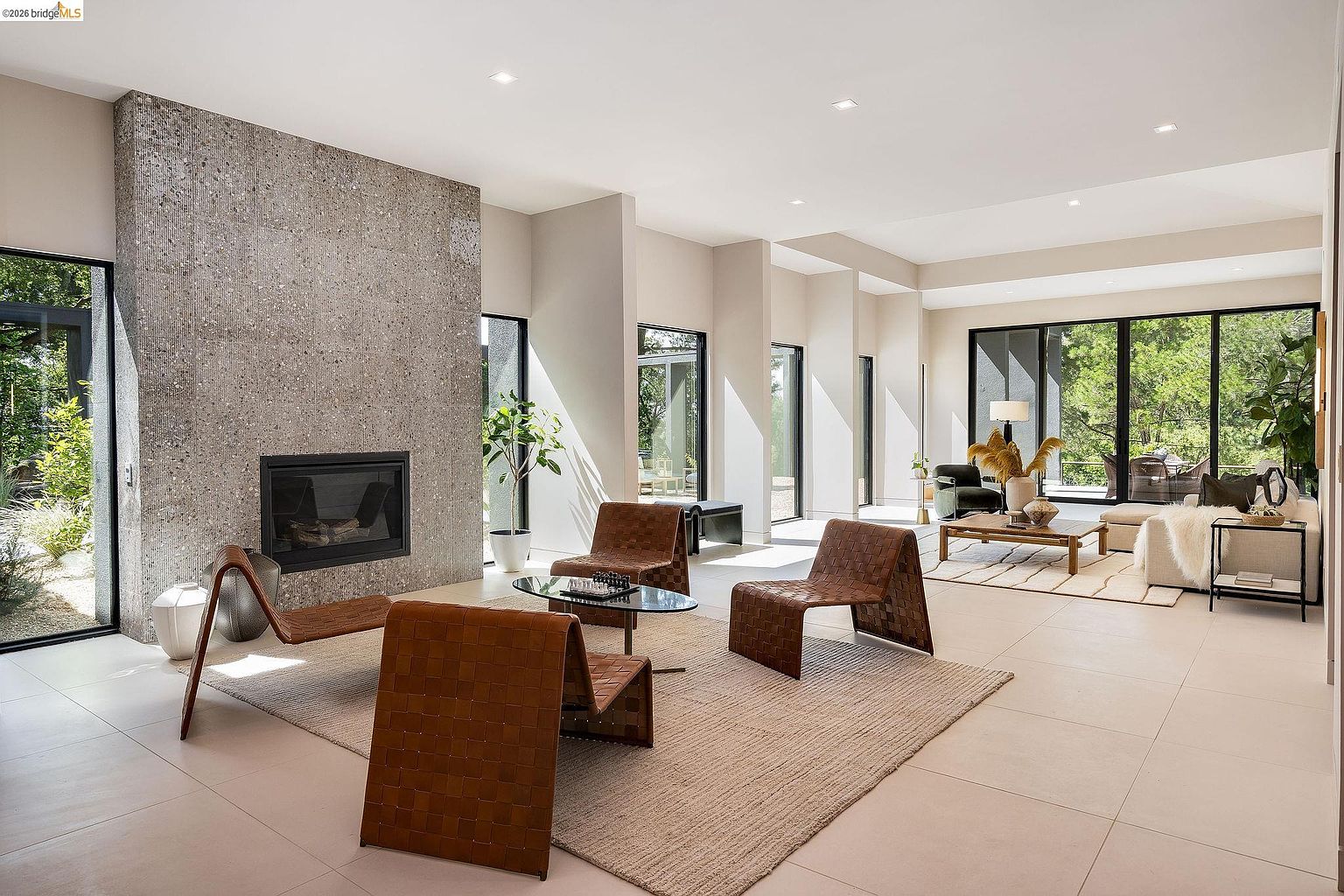 This expansive, modern living room features a striking floor-to-ceiling textured stone fireplace as its central focal point. The open-concept space is bathed in natural light from large glass sliding doors that offer seamless indoor-outdoor flow, complemented by minimalist furniture and neutral-toned tile flooring. The room exudes a sophisticated, airy atmosphere, perfect for high-end contemporary living.