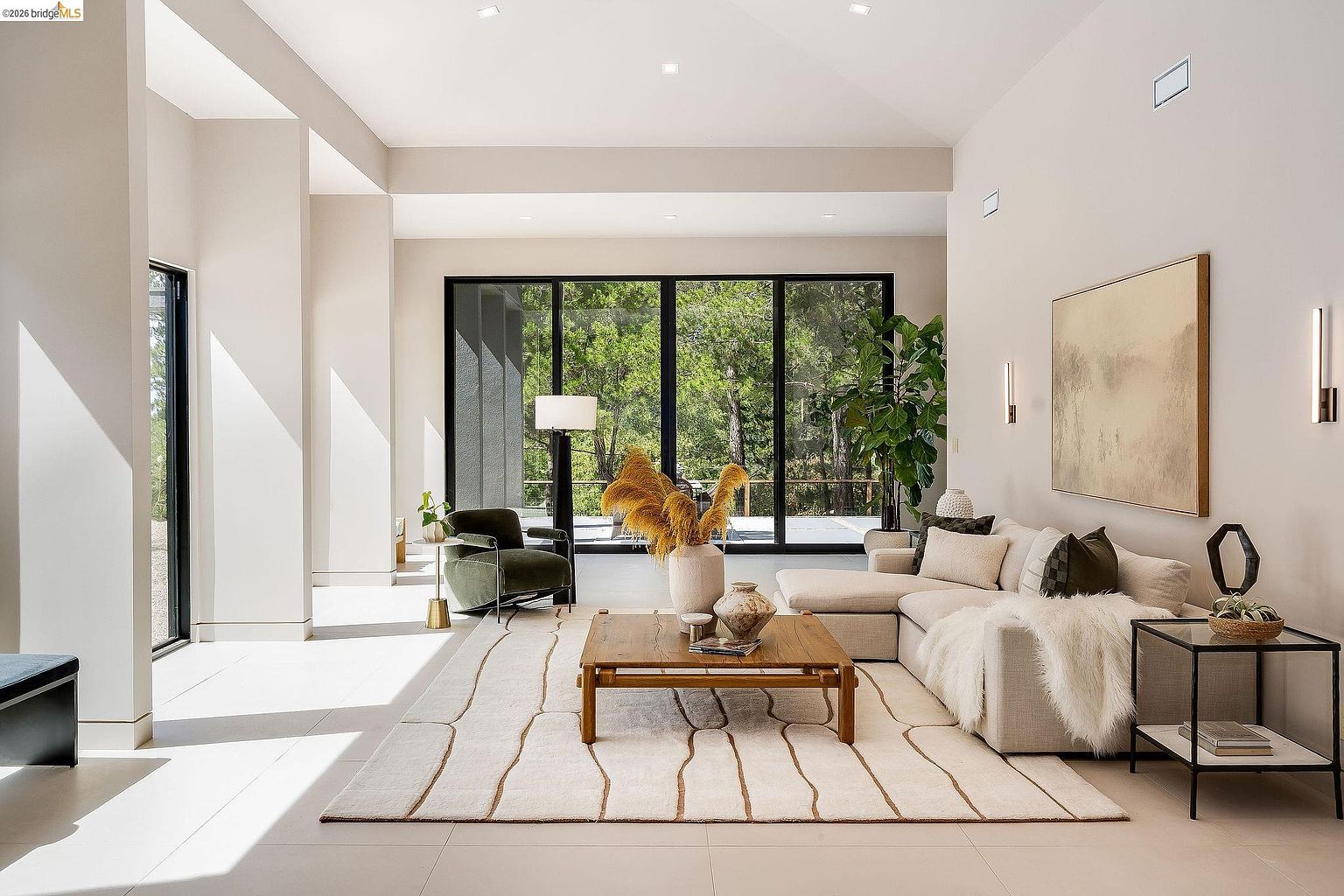 This spacious, modern living room features high ceilings and expansive floor-to-ceiling windows that frame a lush, wooded outdoor view. The room is anchored by a large, light-colored sectional sofa, a rustic wooden coffee table, and a plush area rug, creating a warm and inviting atmosphere. The minimalist design is complemented by elegant wall sconces and a large piece of abstract art, emphasizing a sophisticated and airy aesthetic.