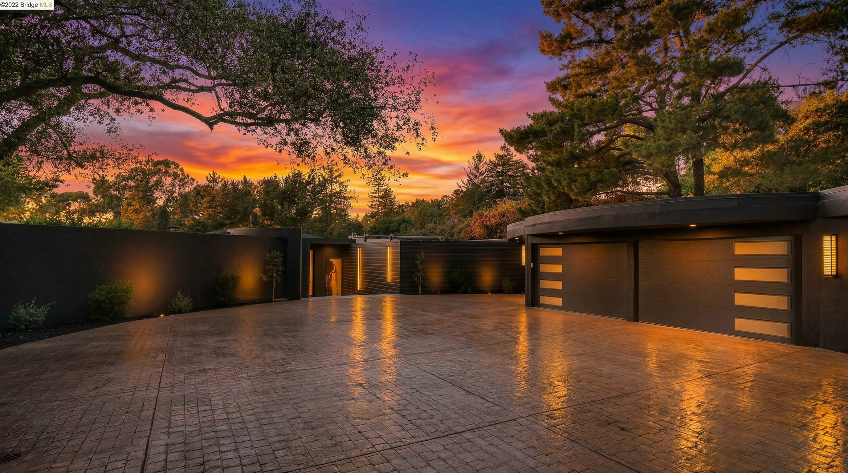 This modern, architectural home showcases a striking dark exterior with a smooth, minimalist aesthetic and integrated linear lighting. The expansive paved driveway leads to a double garage and a grand entryway, set against a dramatic, colorful sunset sky that highlights the property's sophisticated design and curb appeal.
