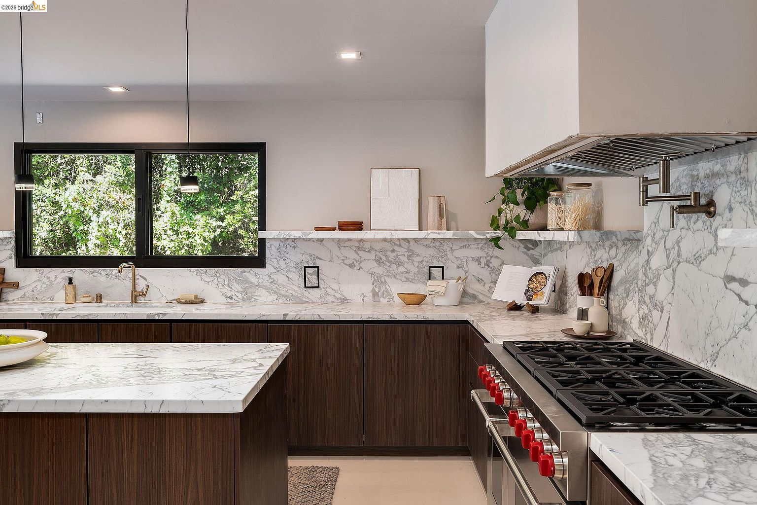 This sophisticated, modern kitchen features rich, dark wood cabinetry contrasted by luxurious white marble countertops and a matching full-height backsplash. A professional-grade gas range with red knobs serves as a focal point, while a large island and floating shelves add both functionality and a sleek, minimalist aesthetic. The space is brightened by natural light streaming through a large window, creating an inviting and high-end culinary environment.