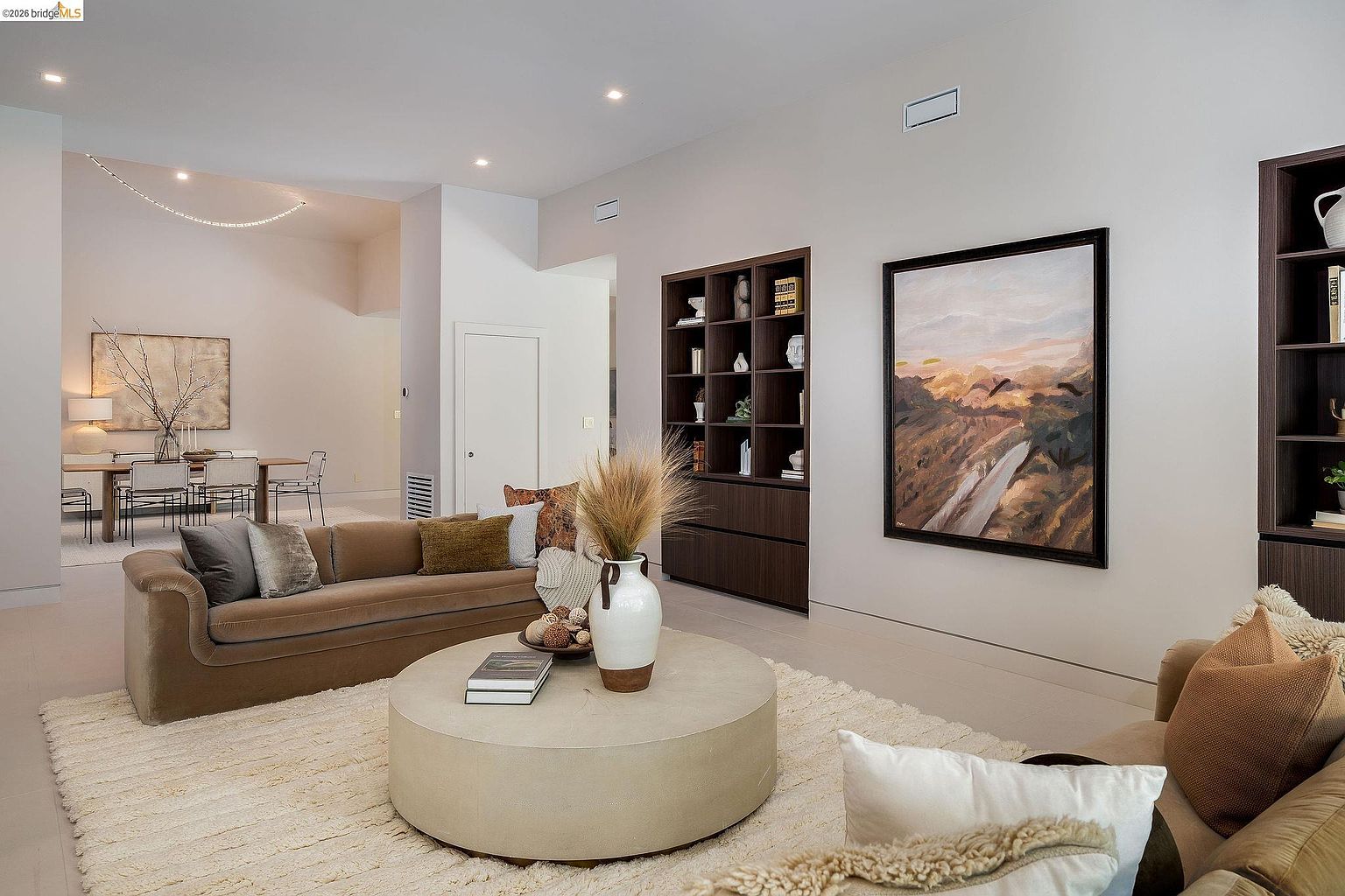 This sophisticated living room features a neutral color palette, a plush cream-colored area rug, and a large, round concrete coffee table as a central focal point. The space is anchored by a sleek, low-profile brown sofa and built-in dark wood shelving, complemented by a large landscape painting that adds warmth to the room. The open-concept layout flows seamlessly into a dining area in the background, creating an airy and inviting atmosphere perfect for modern living.