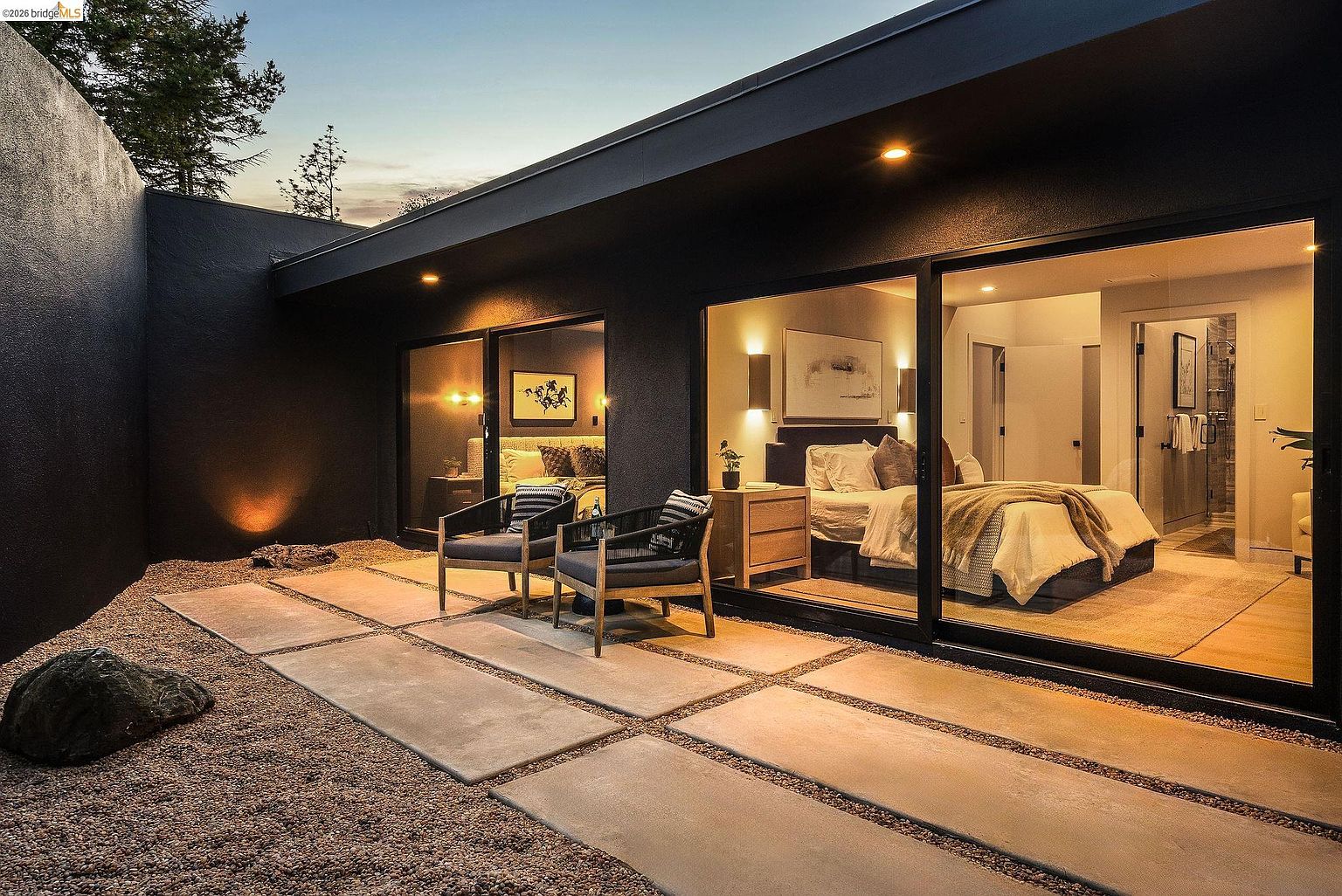 This sophisticated modern patio features large concrete pavers set in a gravel bed, creating a clean, minimalist aesthetic that seamlessly connects to the interior bedrooms through expansive sliding glass doors. The dark exterior walls and warm, recessed lighting provide a dramatic, high-end atmosphere, while two comfortable armchairs offer a perfect spot for relaxation. The perspective is a low-angle, wide shot that emphasizes the architectural integration between the indoor living space and the private outdoor retreat.