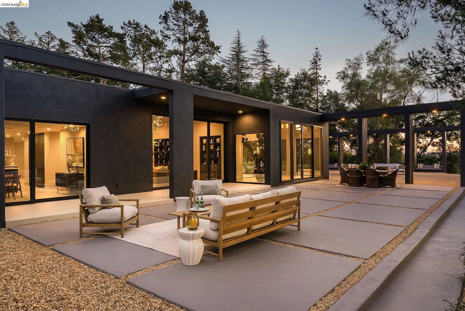 This stunning modern patio features large concrete pavers set in gravel, creating a seamless transition between the indoor living spaces and the outdoor environment. The sleek, dark-painted architectural structure frames the area, which is furnished with comfortable, contemporary outdoor seating and a dining set under a pergola. The scene is captured at dusk, with warm interior and exterior lighting highlighting the sophisticated, minimalist design and the surrounding natural landscape.