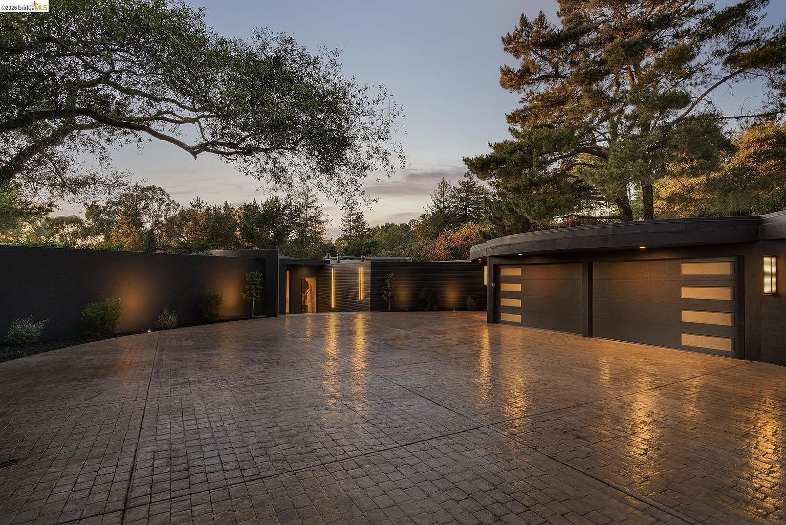 This striking modern residence features a sleek, dark-toned exterior with a curved garage structure and a textured, stamped concrete driveway. The home is illuminated by warm, integrated accent lighting that highlights the architectural lines and creates a sophisticated, inviting atmosphere at dusk. Surrounded by mature trees and a minimalist landscape, the property exudes a sense of luxury, privacy, and contemporary design.